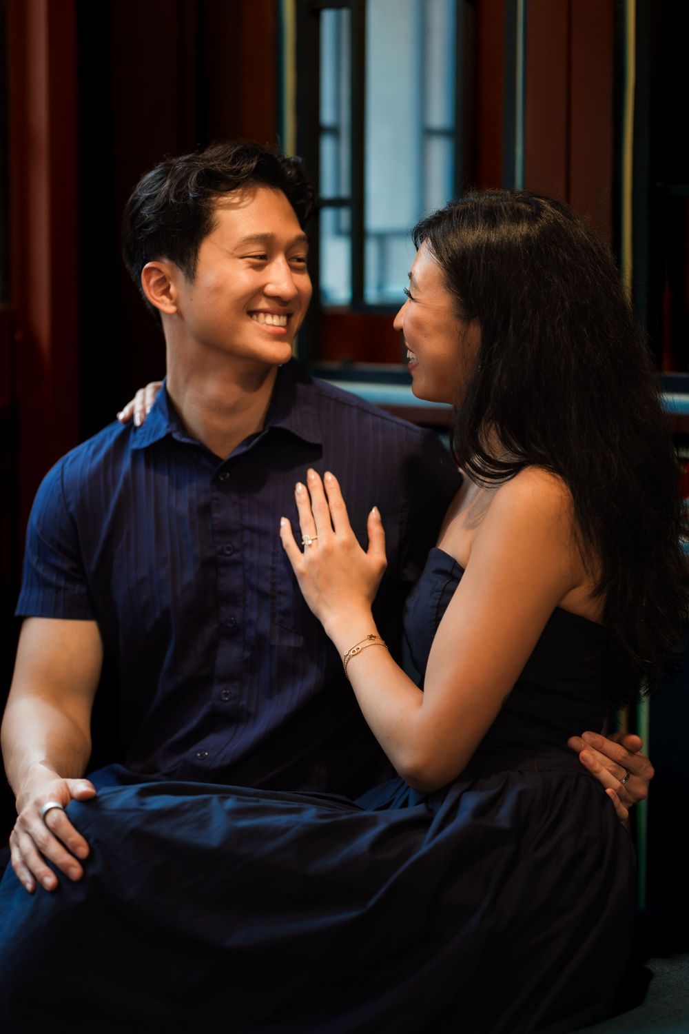 A happy Asian couple in navy attire share an intimate moment indoors, with the woman showing her engagement ring.