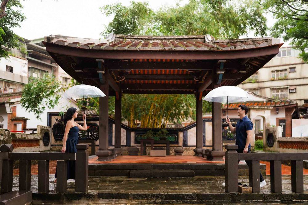 Two people with umbrellas stand beside a traditional Asian wooden pavilion surrounded by bamboo in a rainy courtyard.