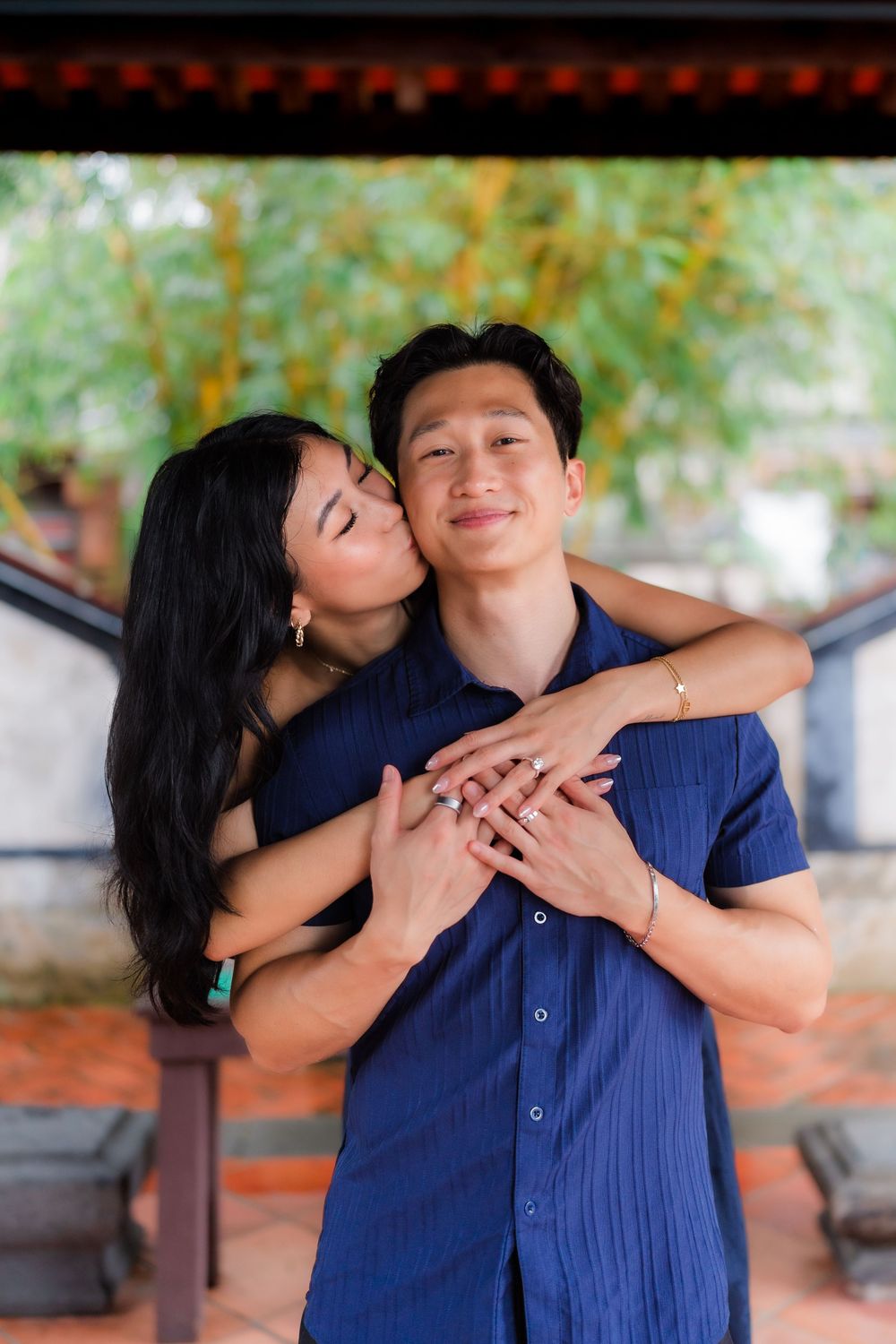 A woman kisses a smiling man on the cheek while embracing him from behind outdoors.