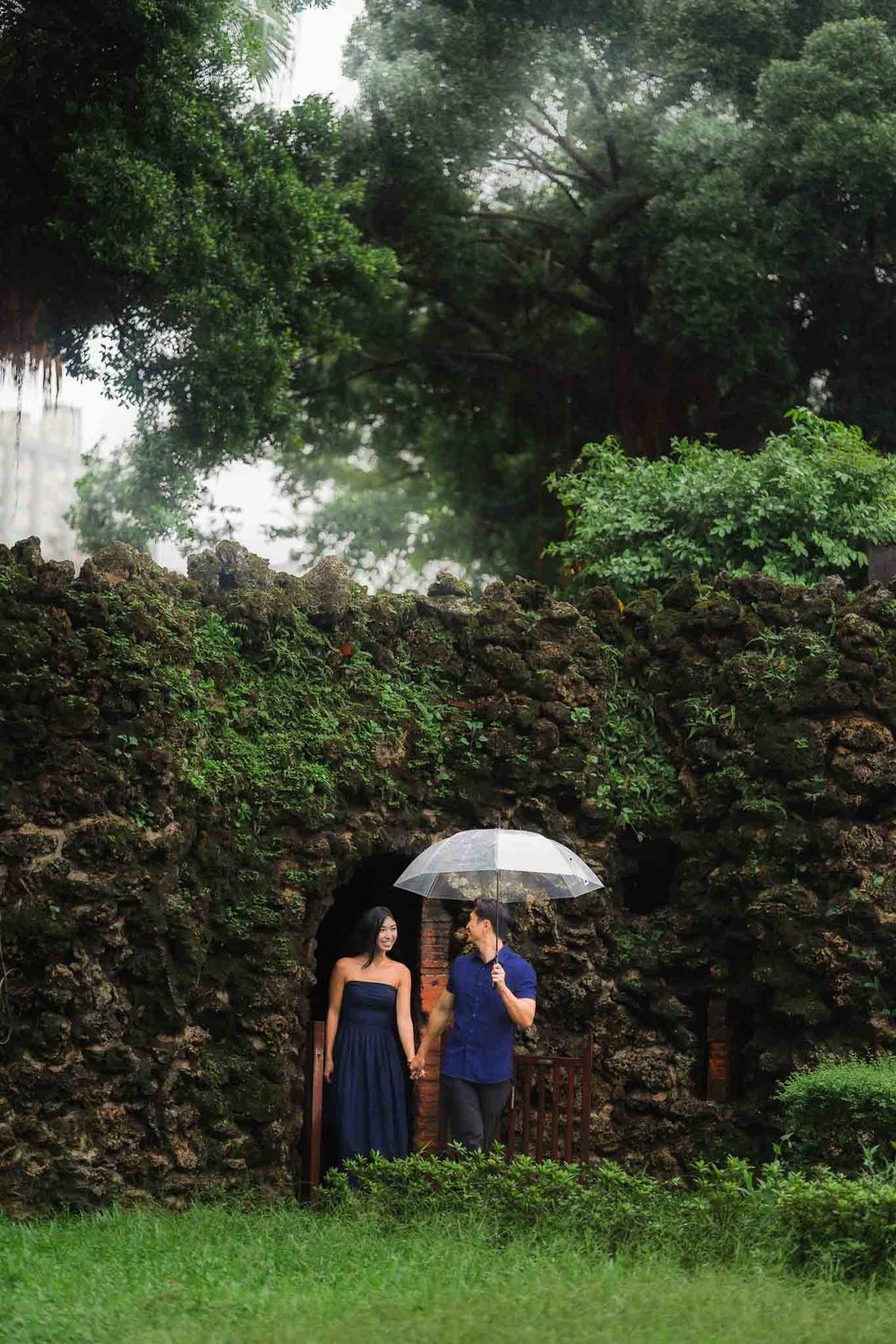 Couple in navy attire sharing umbrella by mossy stone ruins surrounded by lush green trees.