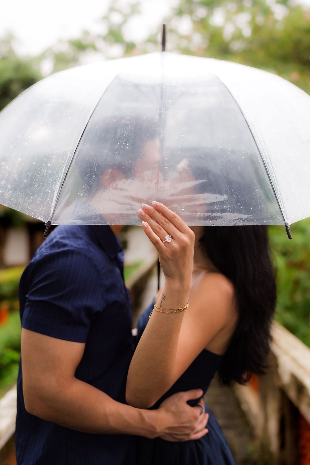 Couple kissing under a clear umbrella in the rain, woman showing engagement ring, romantic outdoor proposal moment.