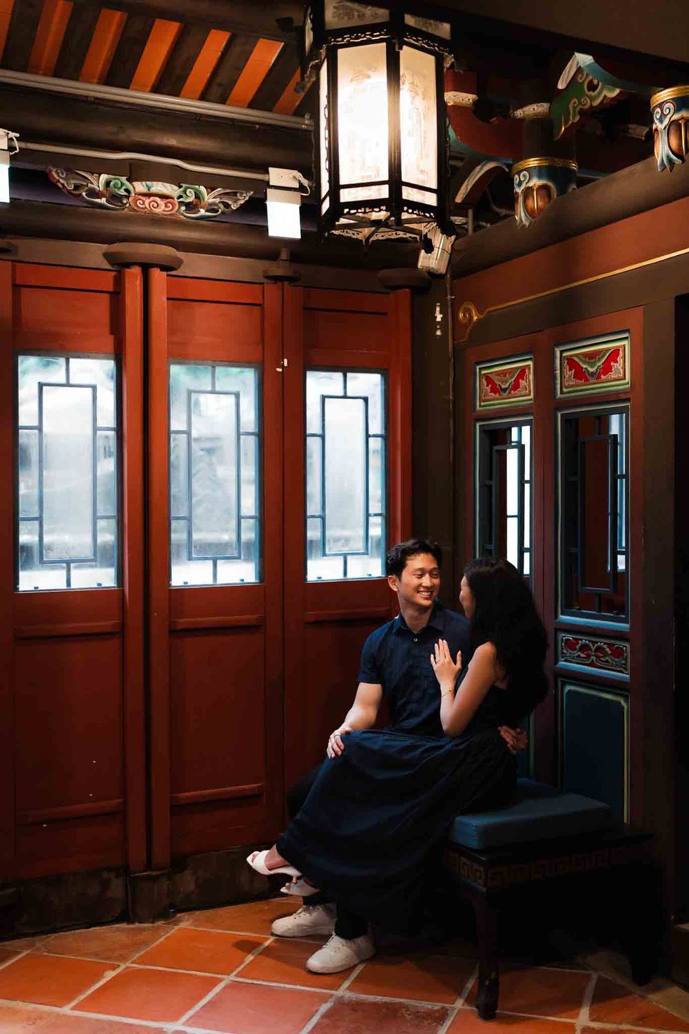 Couple sitting together in a traditional Asian architectural interior with red wooden doors and ornate ceiling lantern.