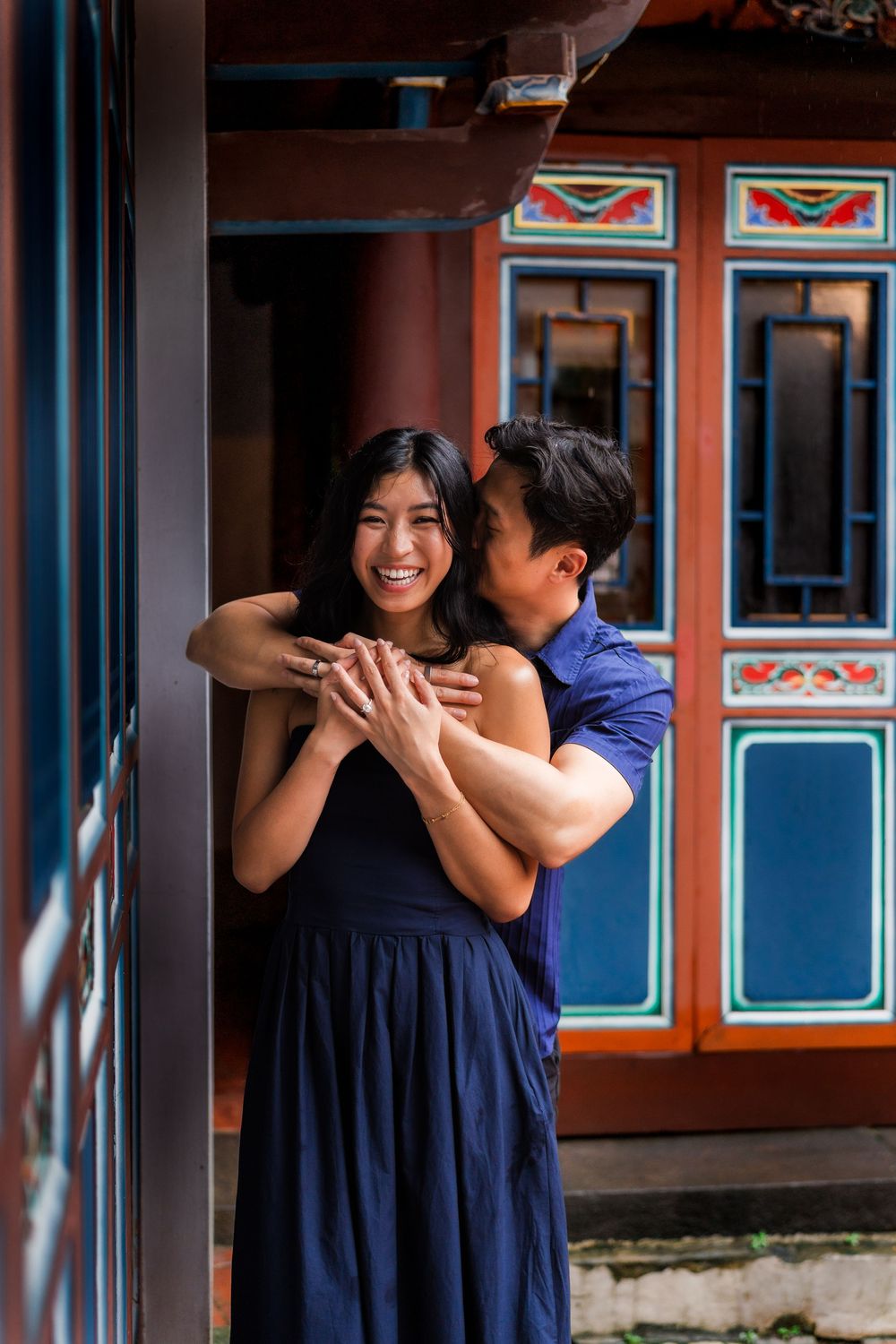 A couple embraces lovingly in front of a colorful traditional Asian building, both wearing navy blue outfits.