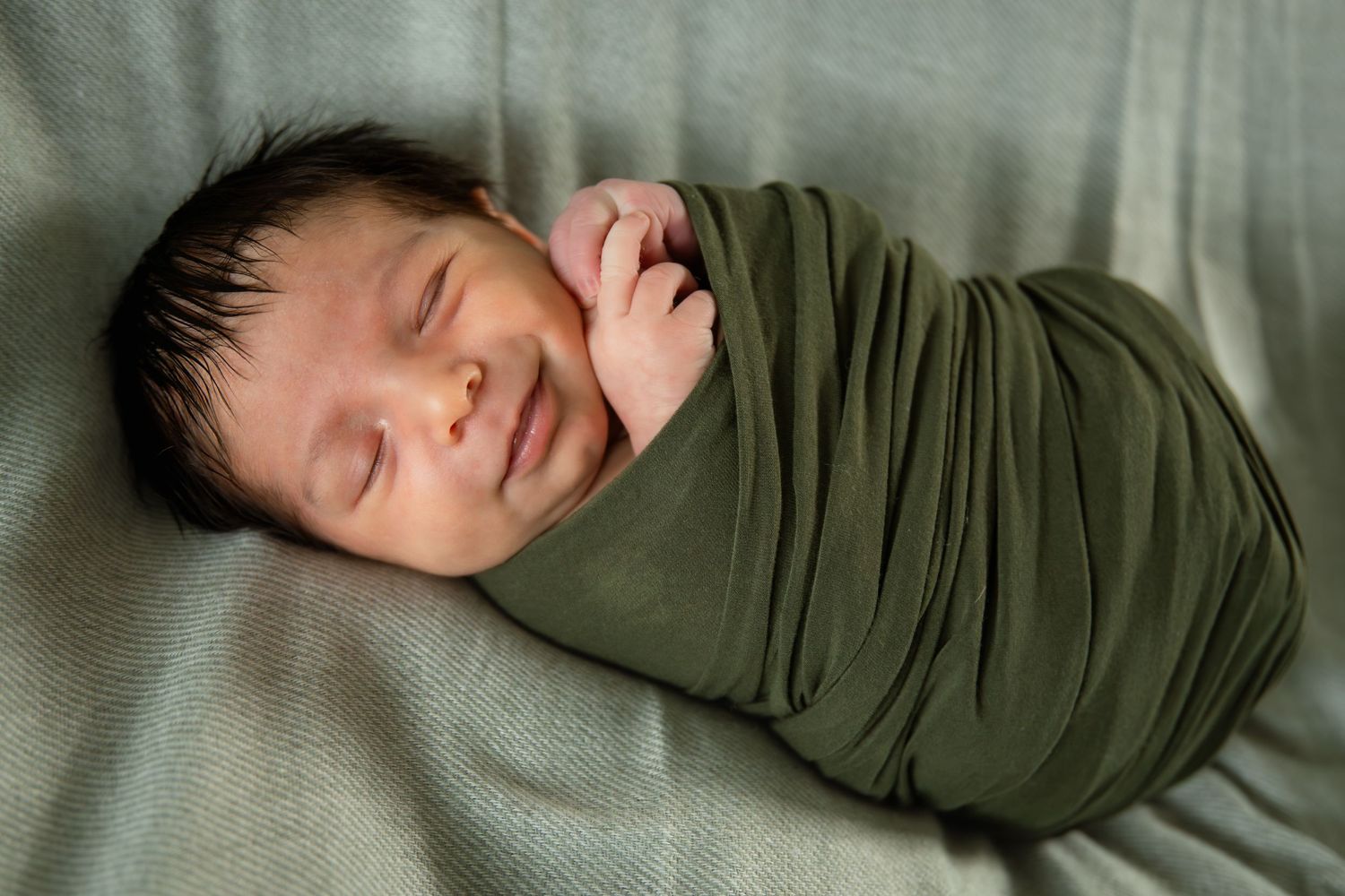 Newborn baby swaddled in olive green wrap, sleeping peacefully with a gentle smile on a gray blanket.