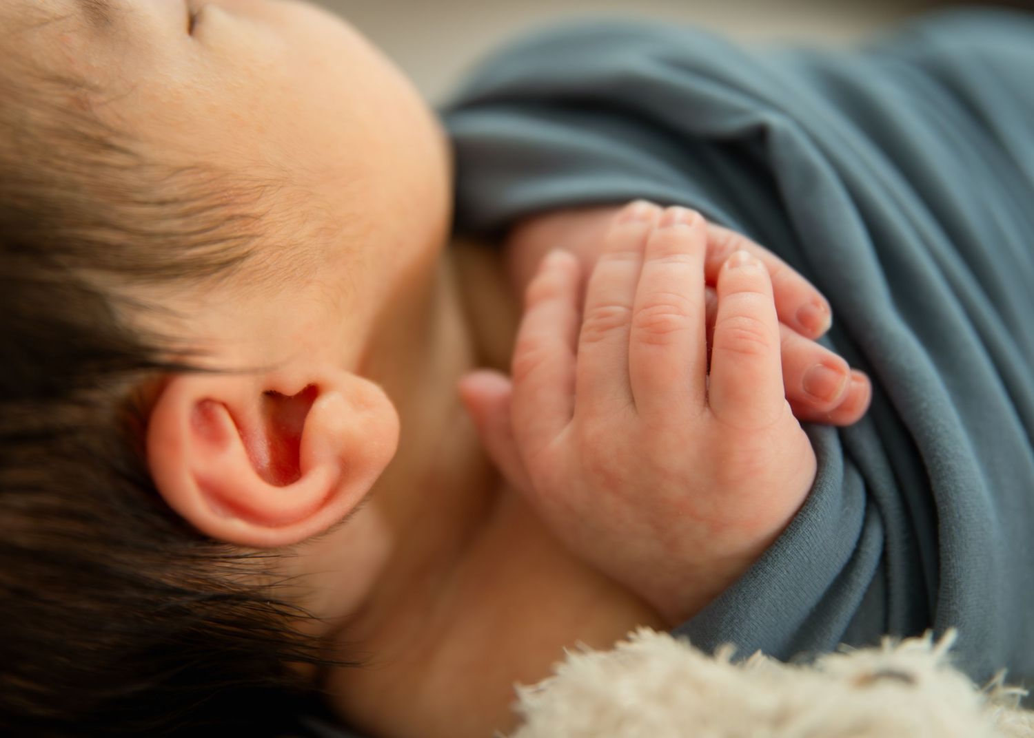 Close-up of a sleeping newborn baby's ear and tiny hand in a blue onesie resting on a soft surface.