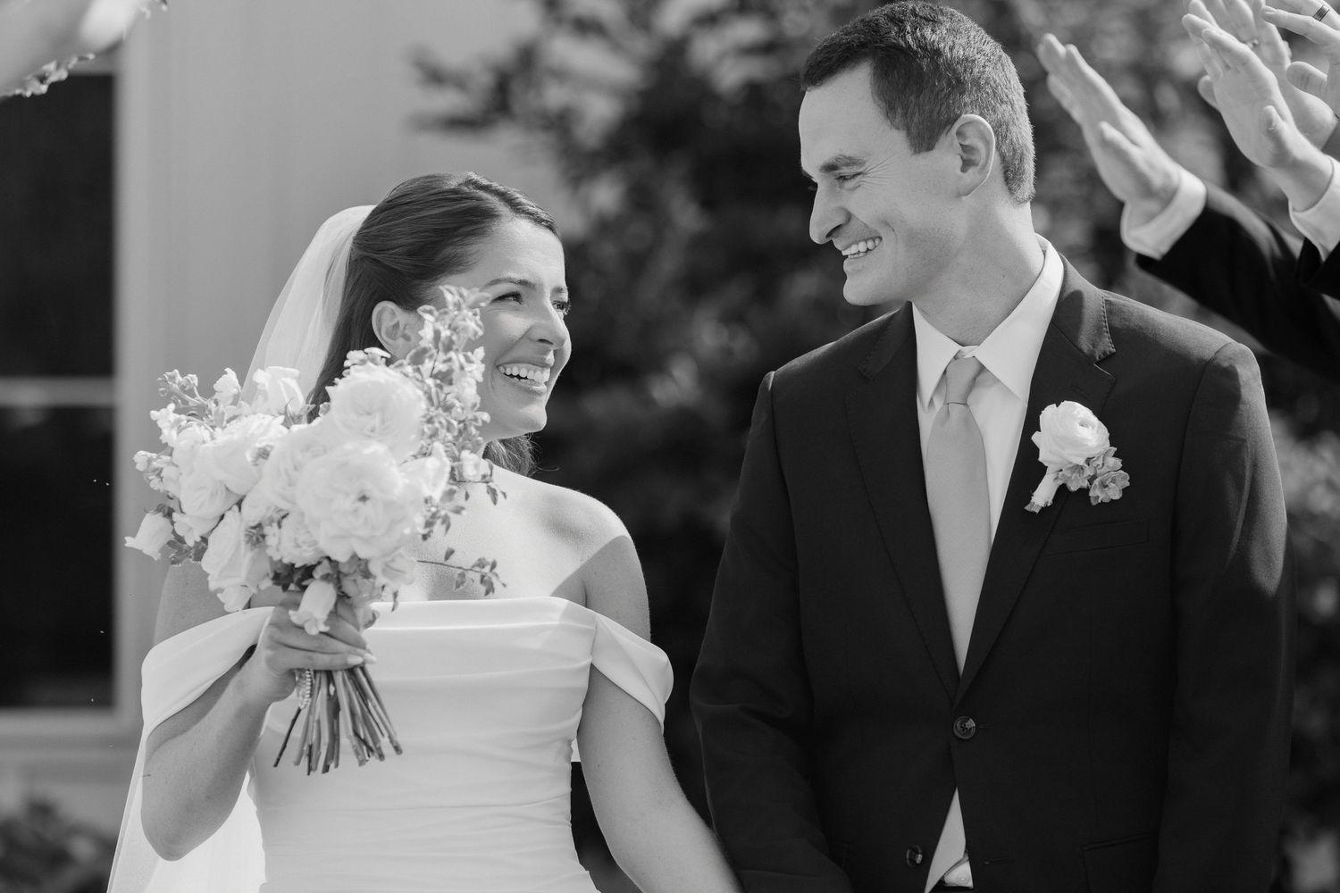 Black and white wedding photo of smiling bride holding bouquet and groom in suit sharing a joyful moment.