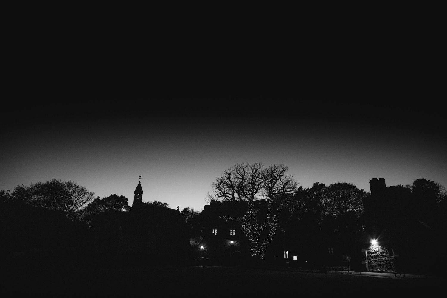 Black and white night scene at Peckforton Castle with the chapel steeple, bare trees and glowing windows silhouetted against a dark sky.