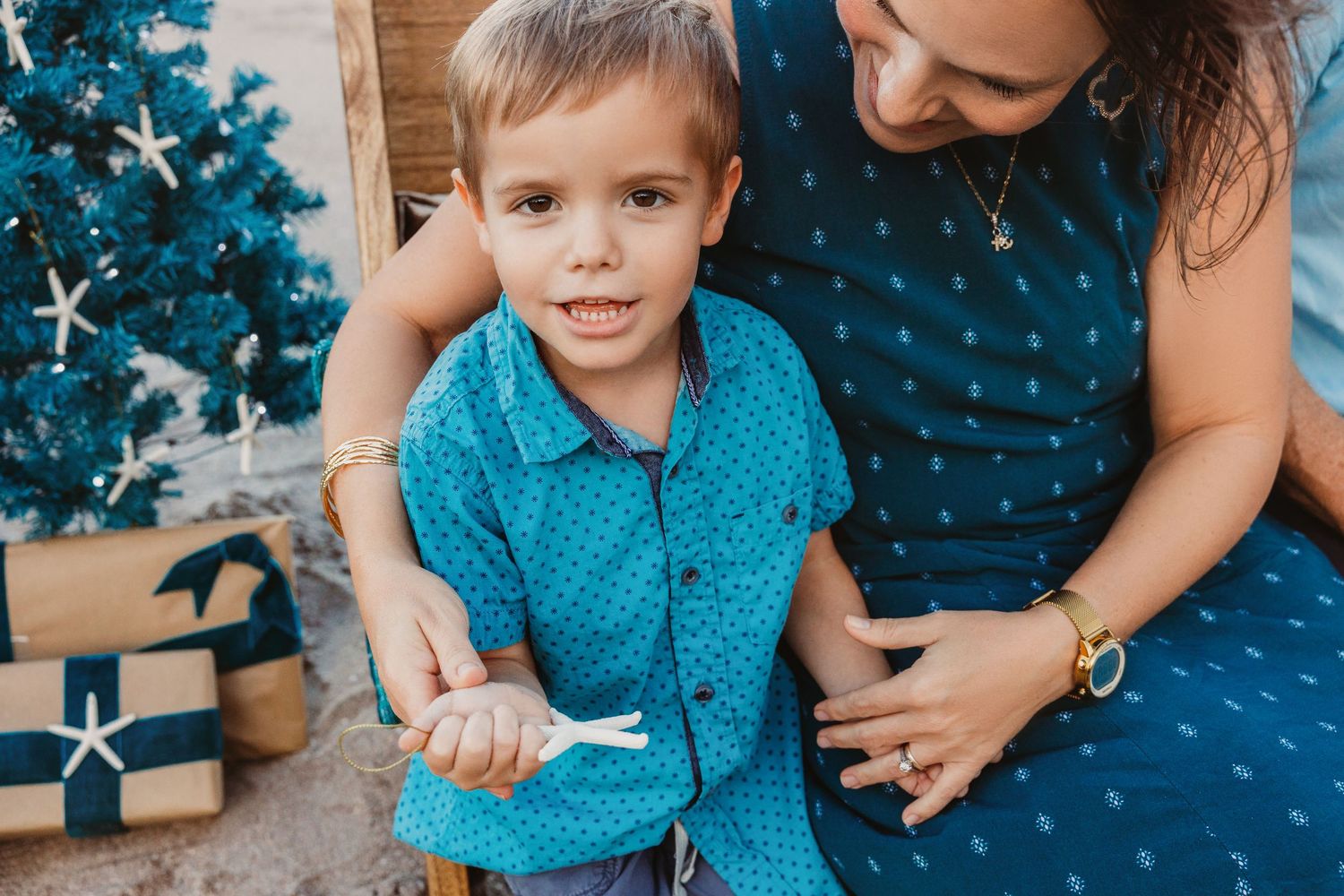 Smiling little boy in a teal button-up shirt sits on a woman's lap beside a decorated Christmas tree and wrapped holiday gifts.