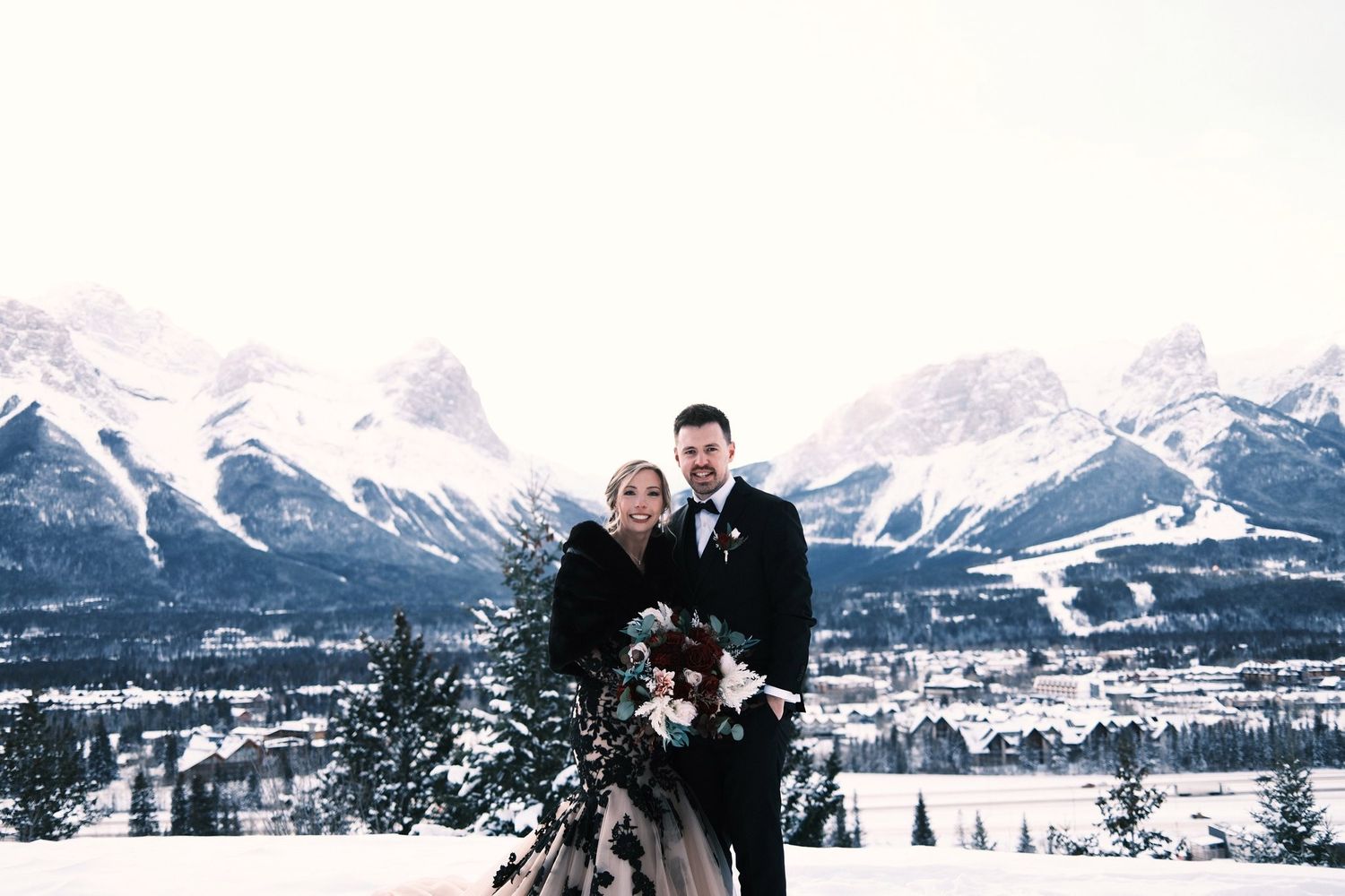 Bride and groom smiling above the Canmore valley with snowy Rocky Mountain peaks and a big winter sky behind them