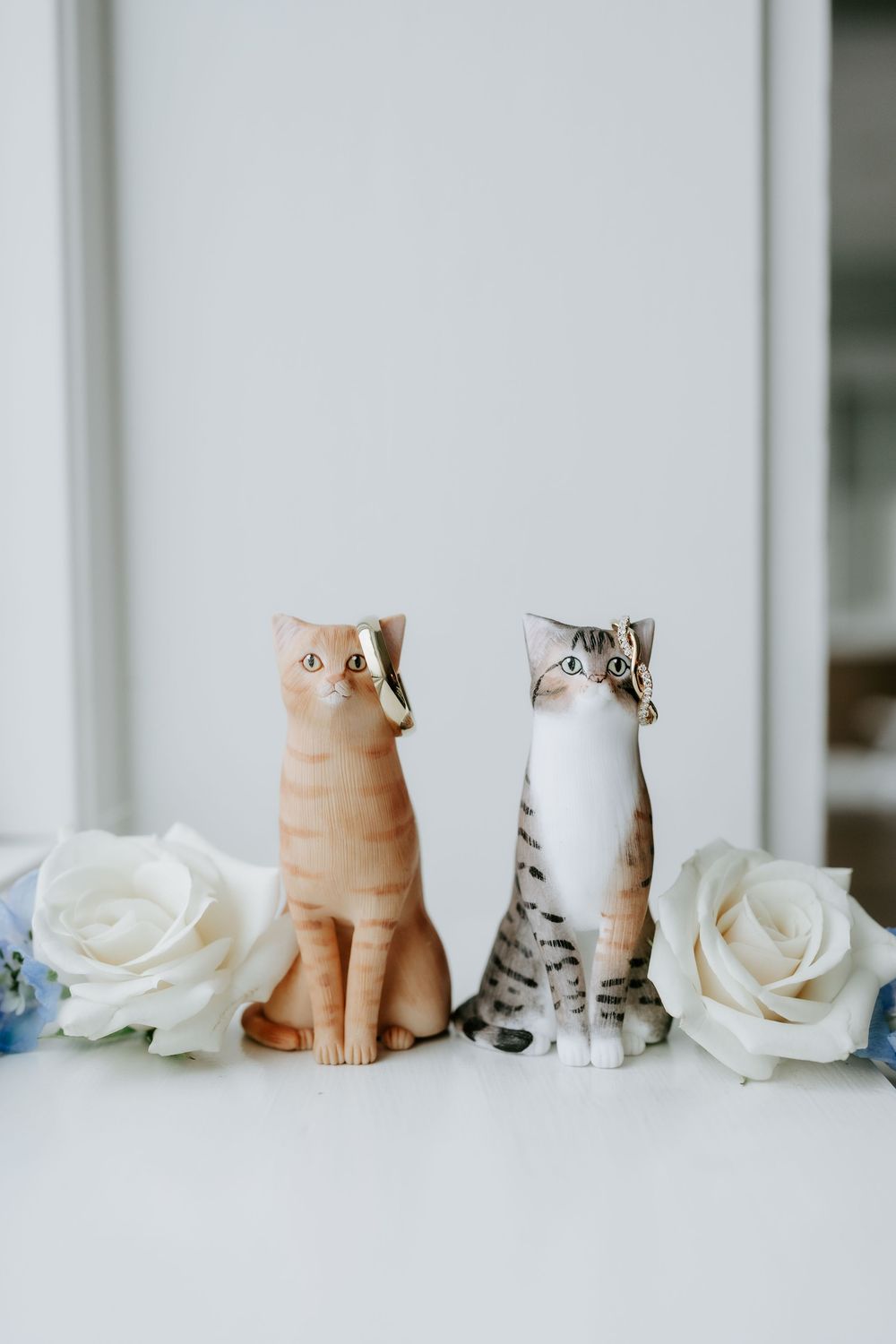 Two ceramic cat figurines with bow ties surrounded by white roses, perfect as wedding cake toppers.