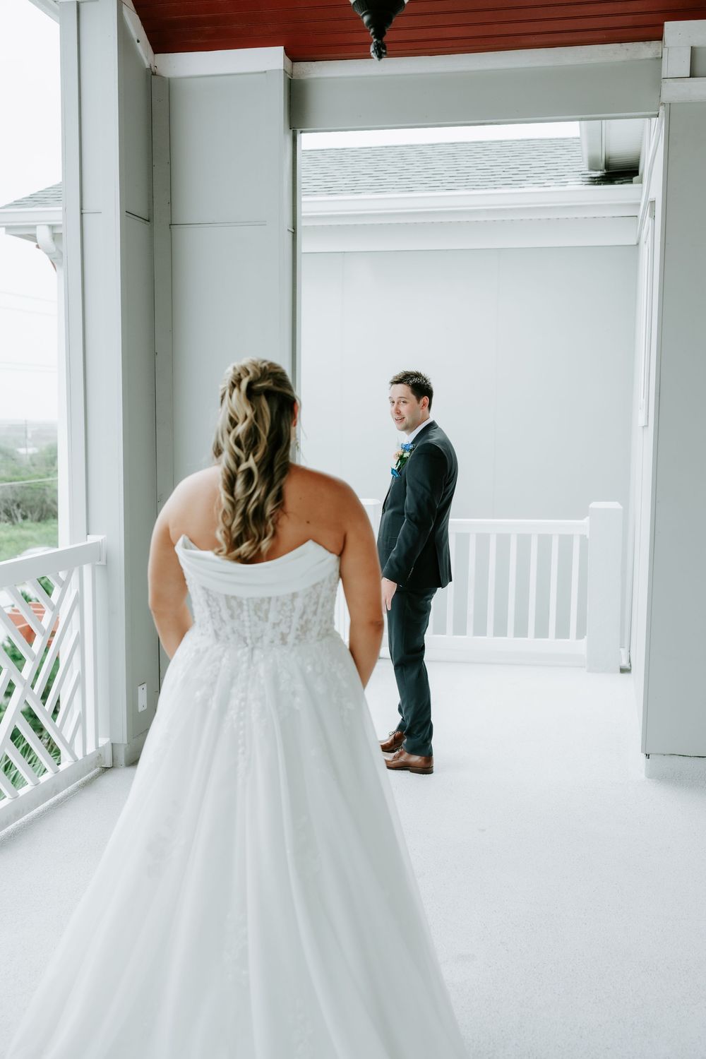 Bride in strapless lace gown faces groom in navy suit during first look on white porch.