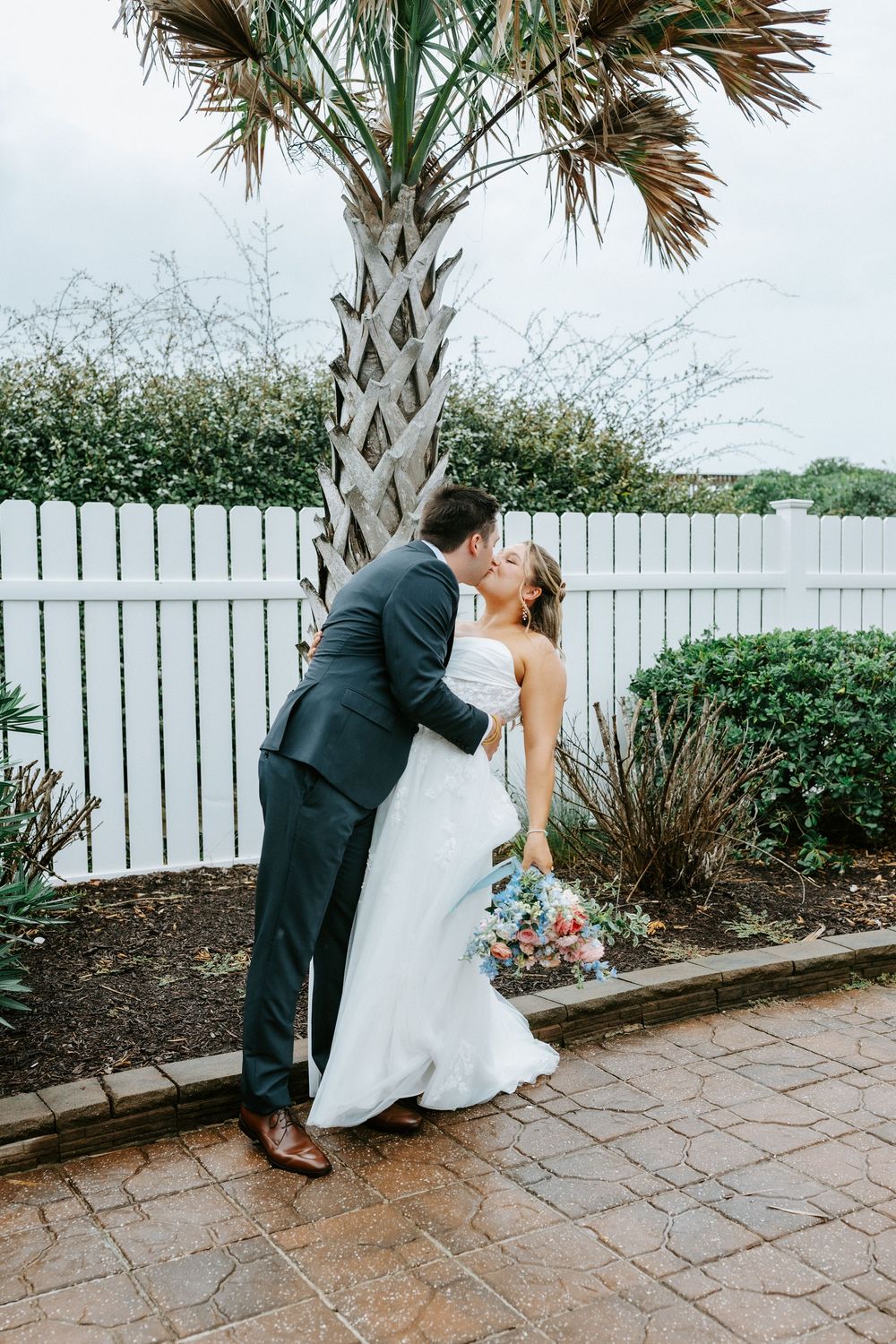 Bride and groom share a romantic kiss under a palm tree beside a white picket fence on their wedding day.