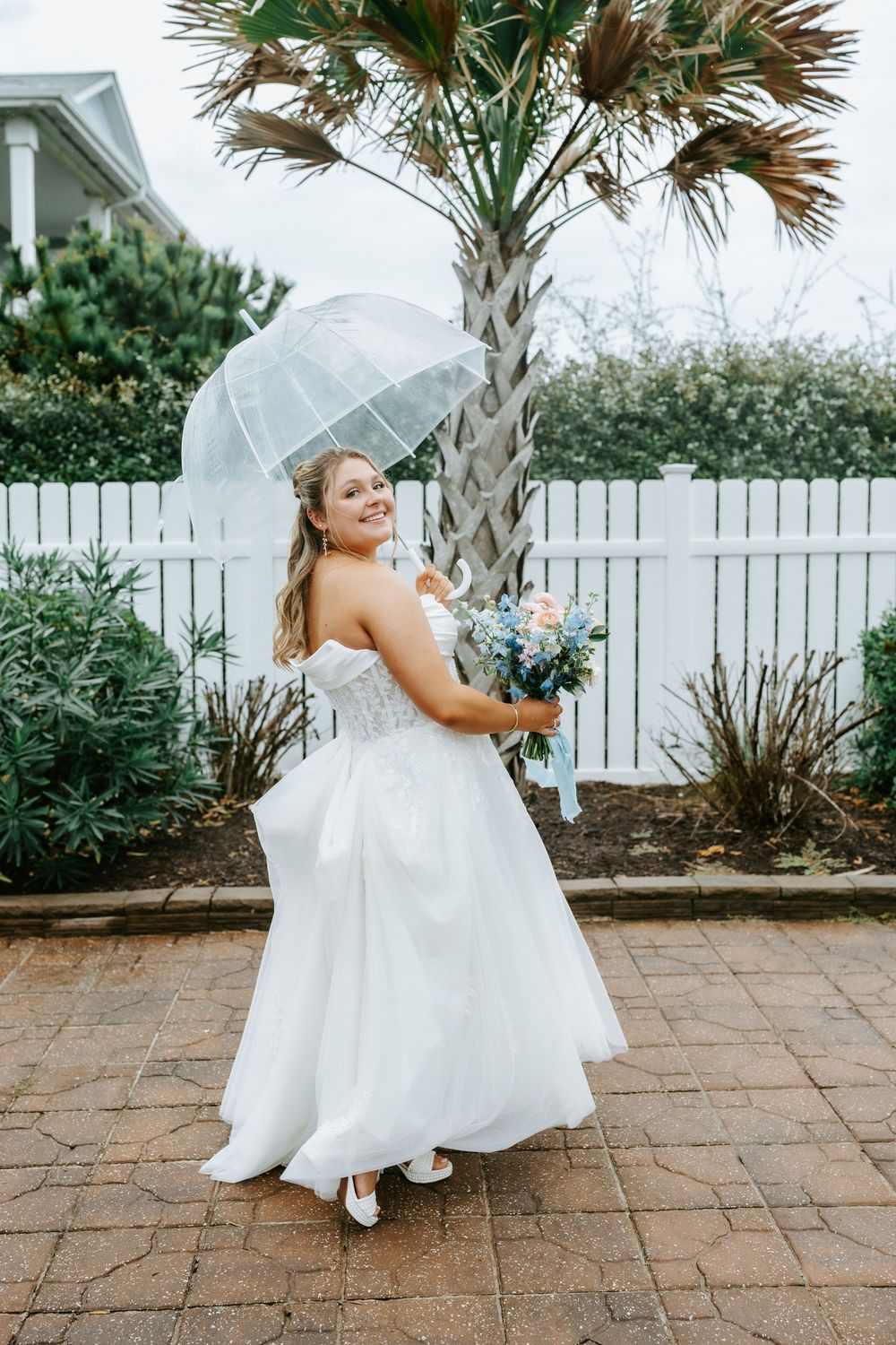 Bride in white strapless gown holds bouquet and clear umbrella on rainy wedding day near palm tree.