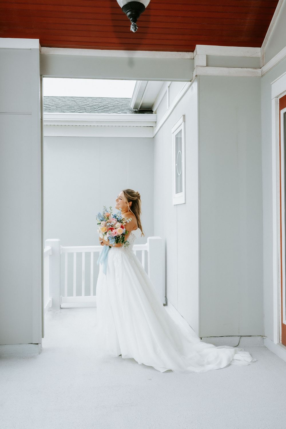 Bride in white gown with floral bouquet standing on porch with red ceiling and white railings.
