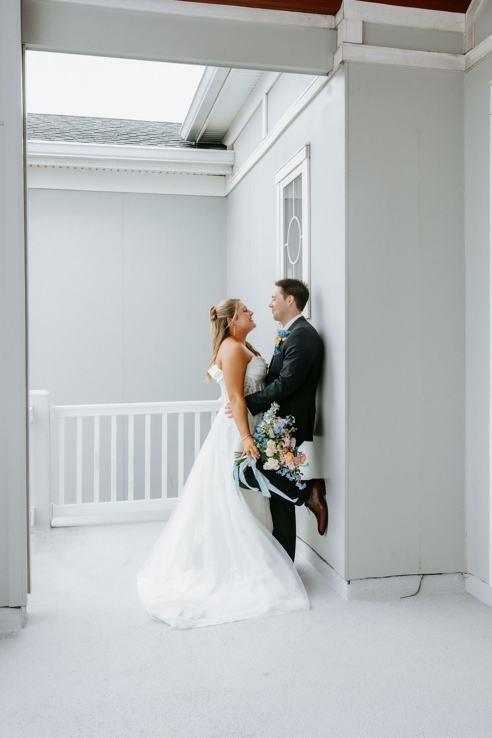 Bride in white gown and groom in dark suit share a romantic moment on a light-filled porch, bride holding a floral bouquet.