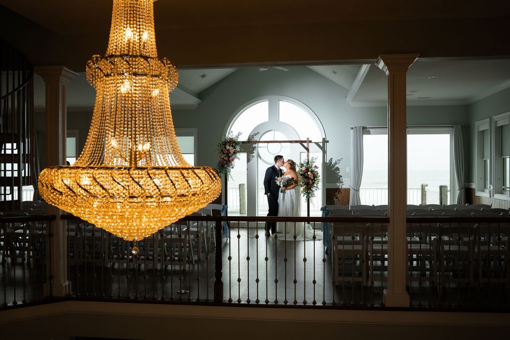 Bride and groom share a romantic moment beneath an elegant gold chandelier in a grand wedding venue.