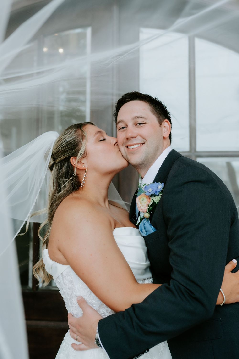 Bride kisses groom on cheek as he smiles, both in wedding attire with floral boutonniere.