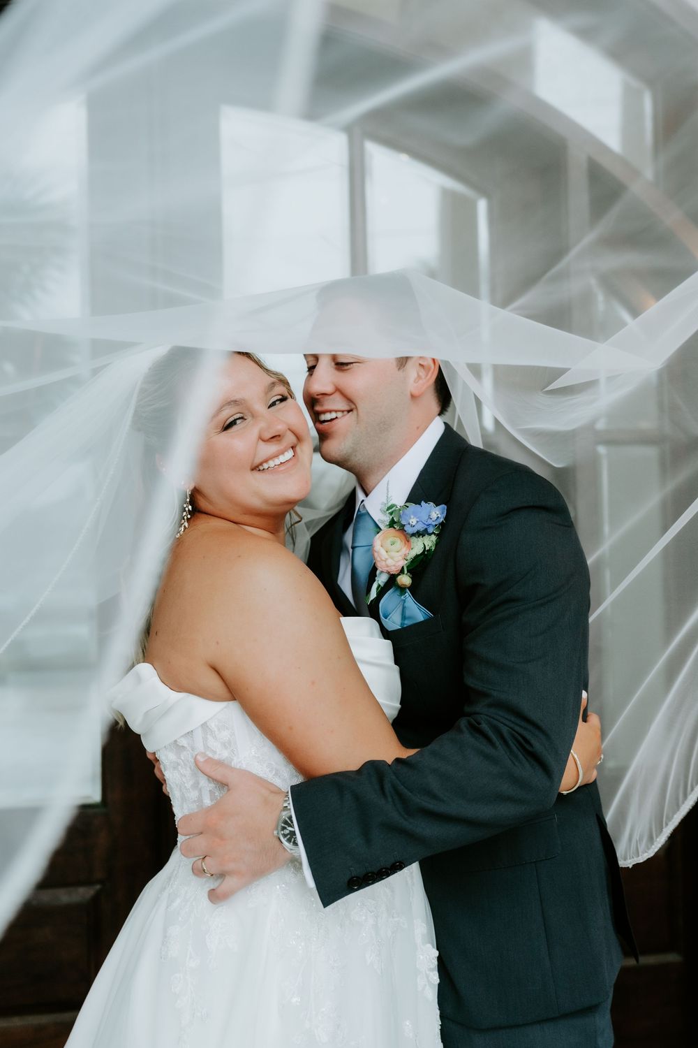 Bride and groom embrace under flowing white veil, smiling joyfully during wedding portrait session.
