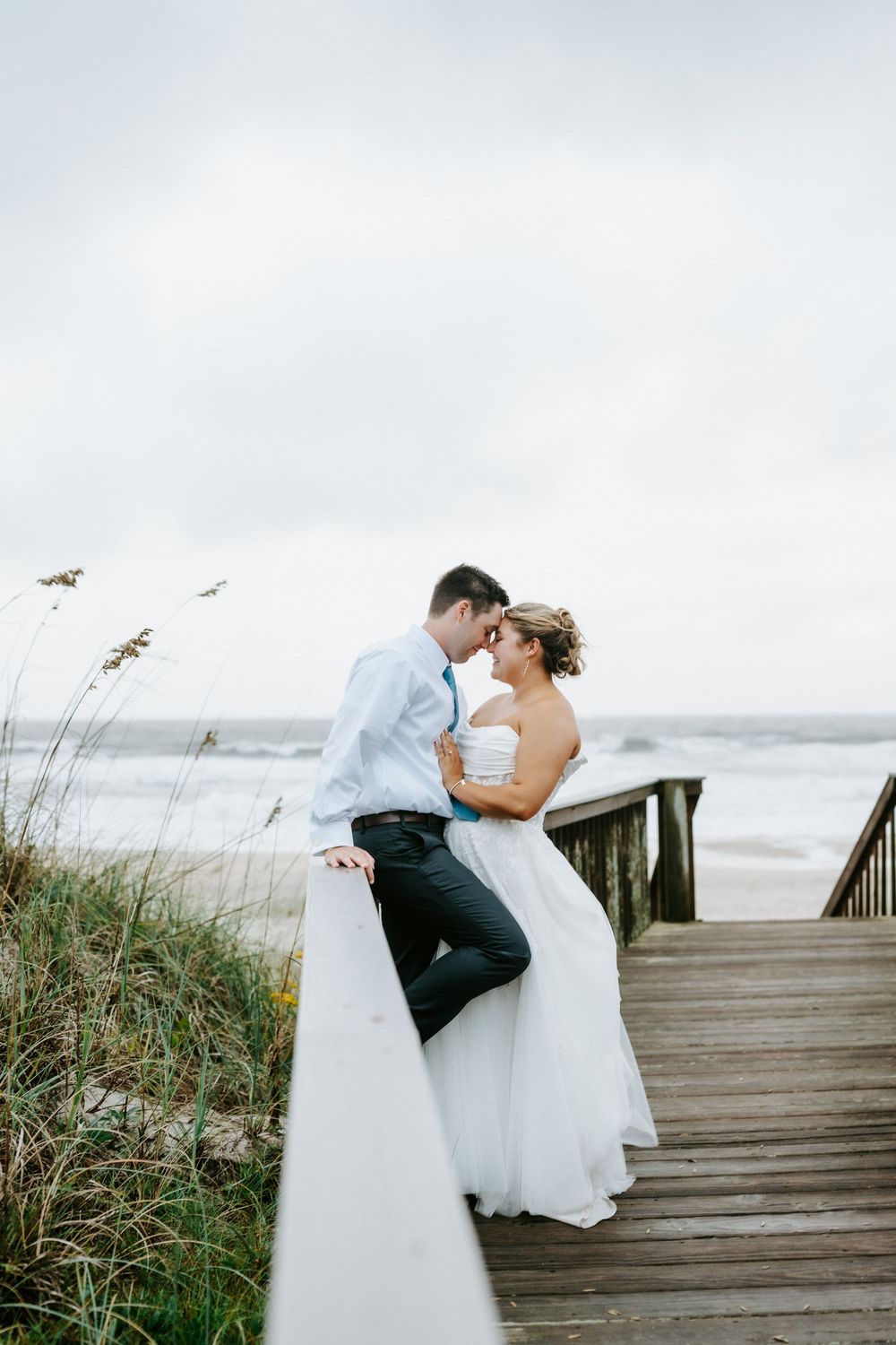 Bride and groom share an intimate moment on a beach boardwalk with ocean waves in the background.