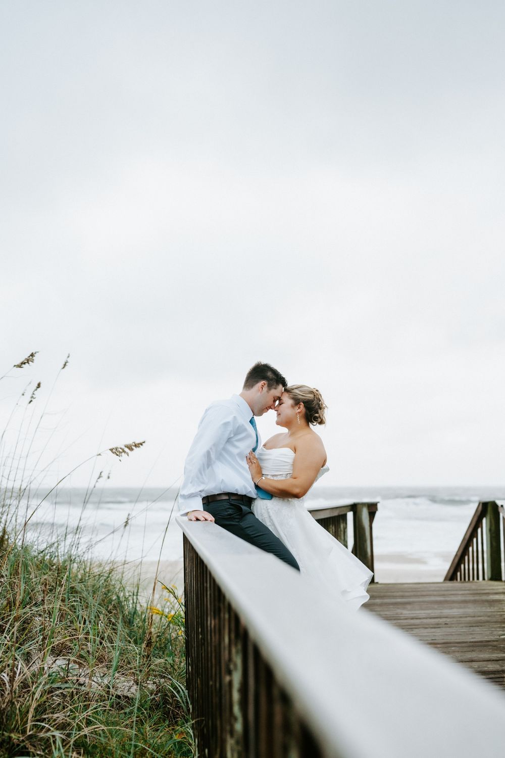 Bride and groom share a romantic moment on a beachside boardwalk, with ocean waves and sea oats in the background.