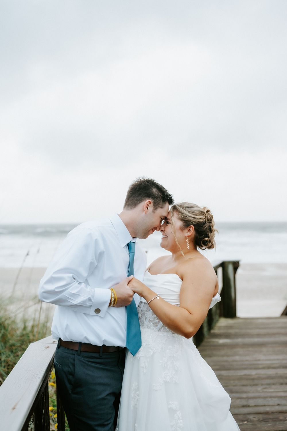 Bride and groom touch foreheads romantically on beach boardwalk under cloudy skies in wedding attire.