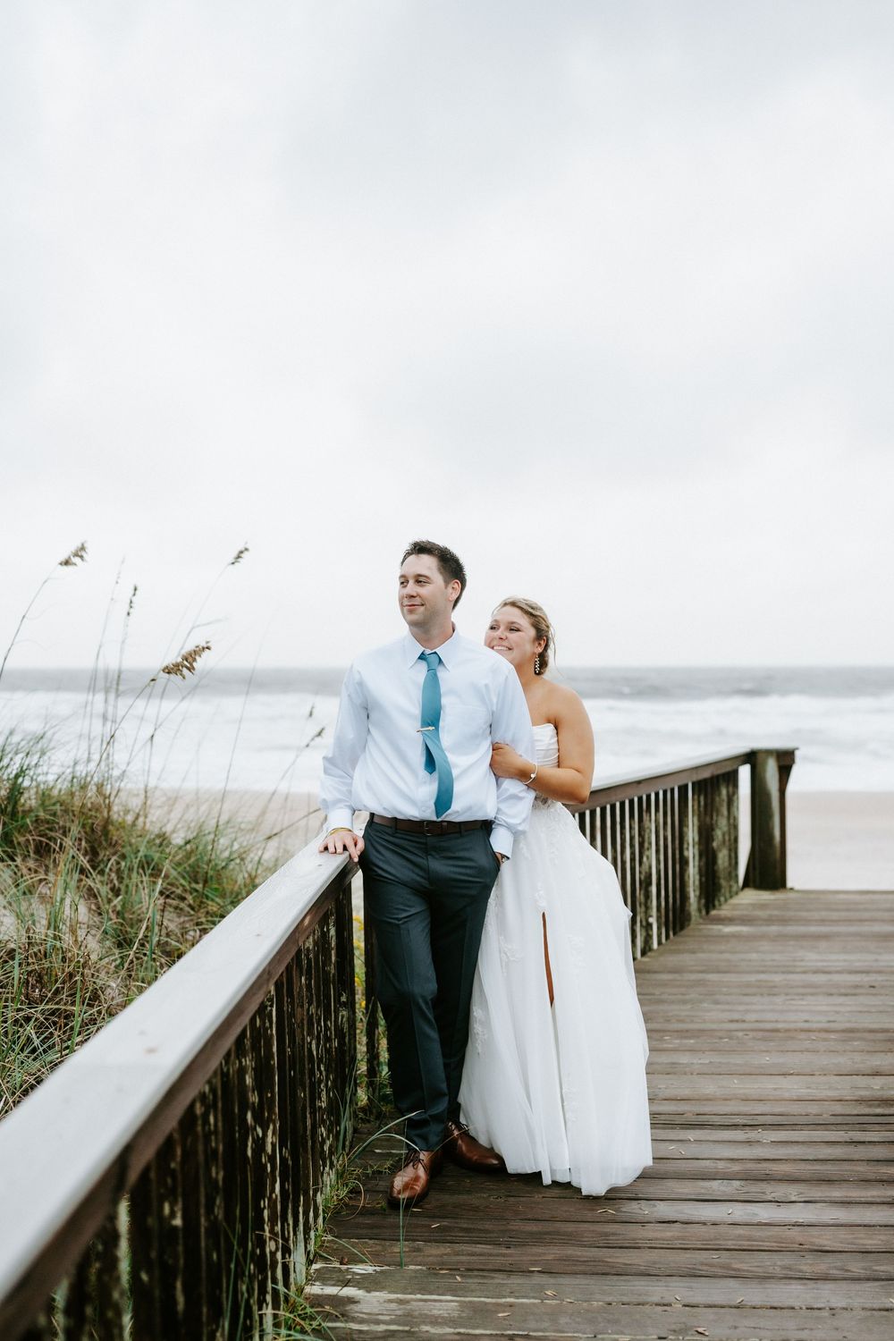 Bride and groom embracing on a wooden boardwalk overlooking a stormy ocean beach on their wedding day.