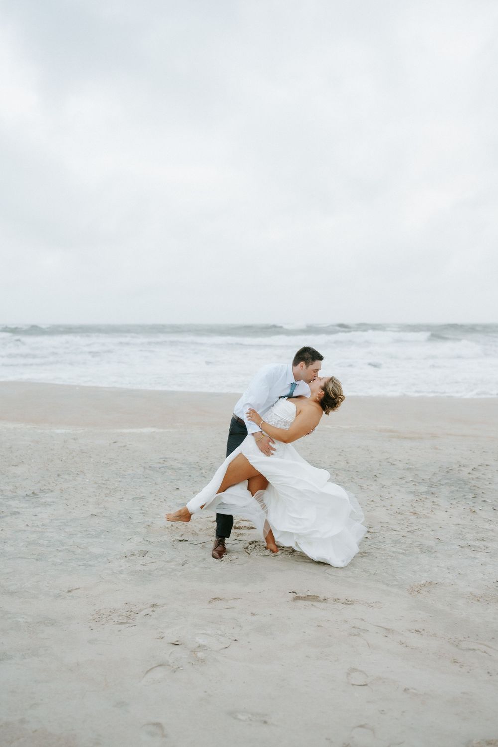 Groom dips bride for a kiss on a sandy beach with ocean waves crashing behind them.