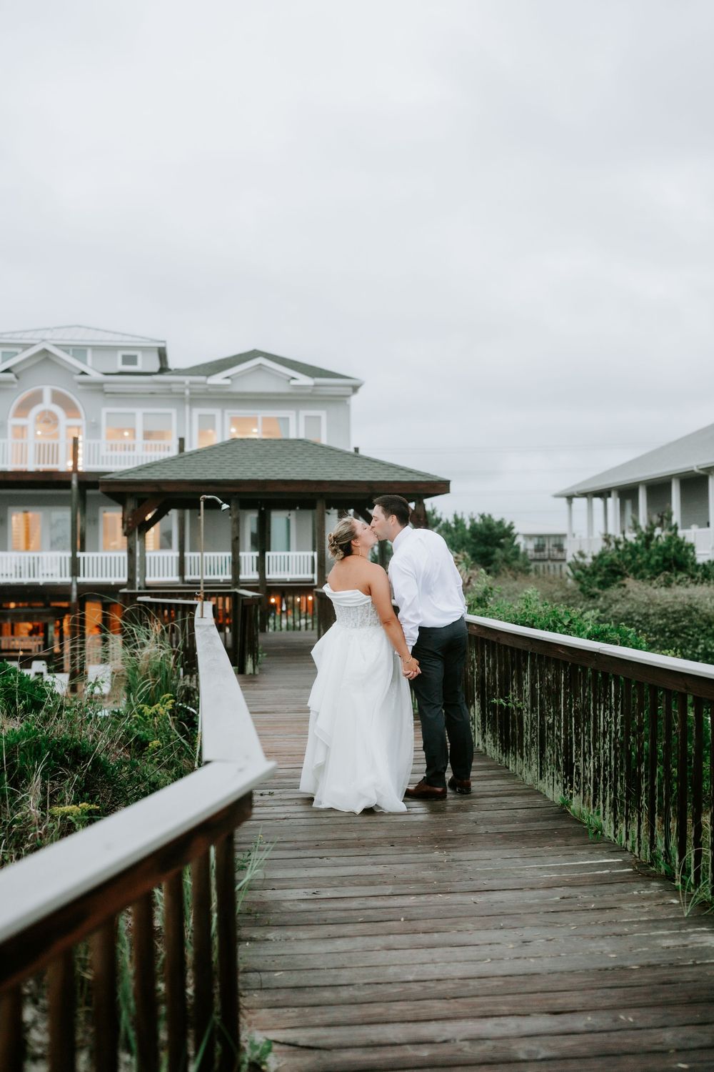 Bride and groom share a kiss on a wooden boardwalk near a coastal beach house on their wedding day.