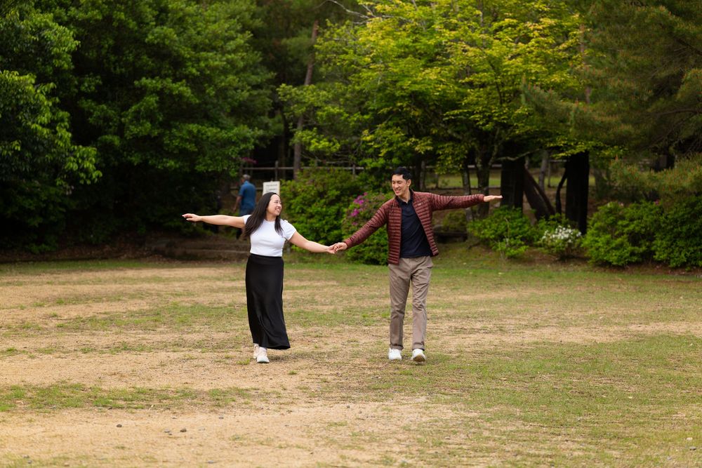 Smiling couple holding hands and dancing on a grassy lawn surrounded by green trees in a sunny park.
