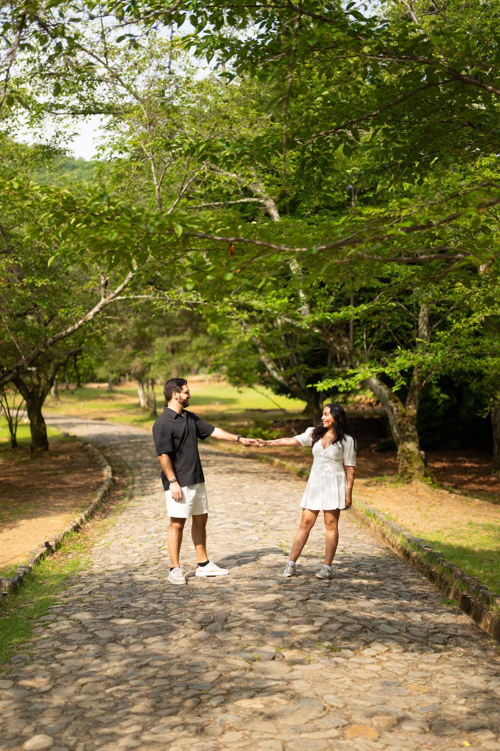 Couple holding hands and smiling at each other on a cobblestone path surrounded by lush green trees in a sunny park.
