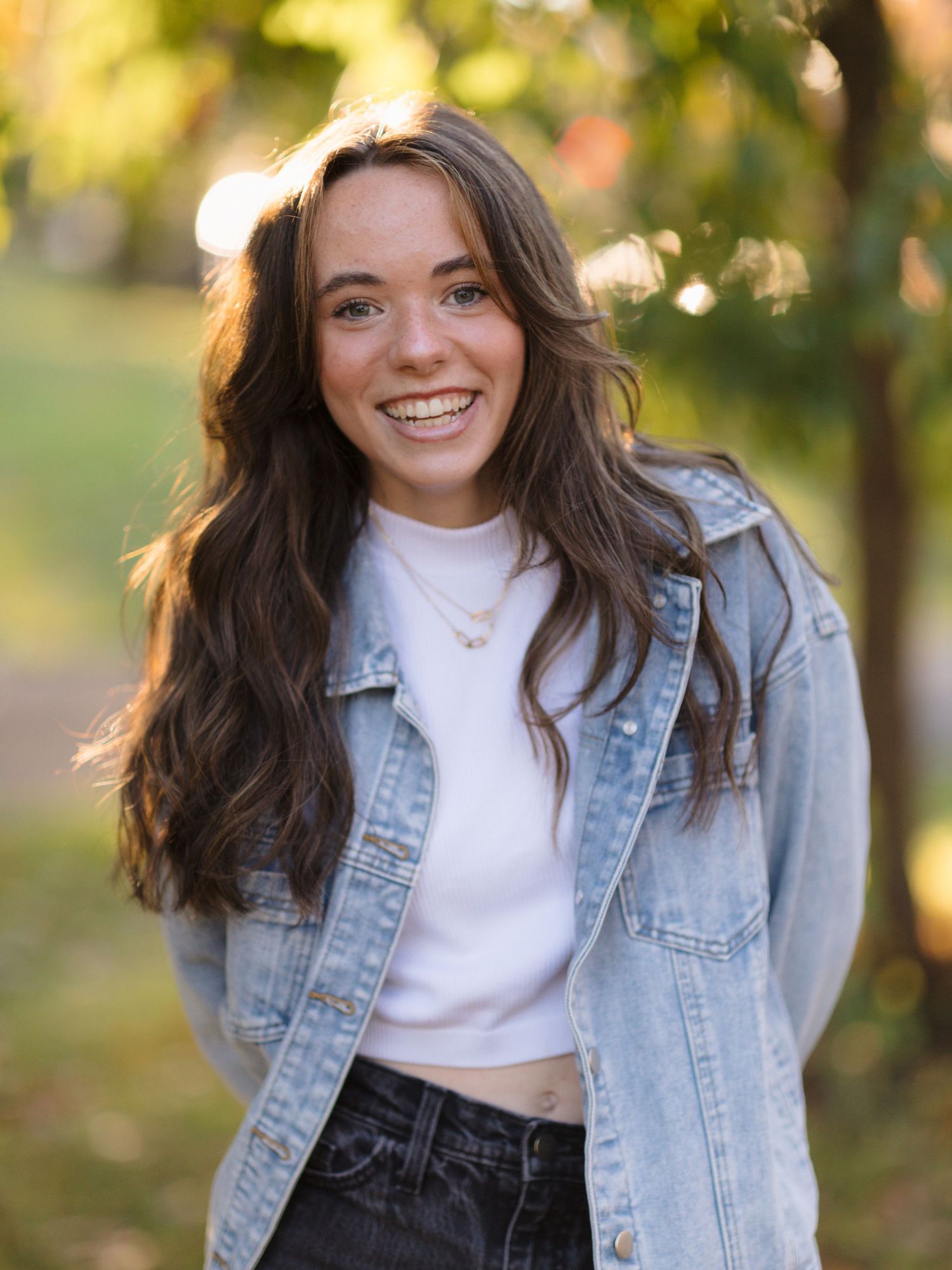 Smiling young woman with long wavy brown hair wearing a denim jacket over a white top, standing outdoors among blurred autumn trees.