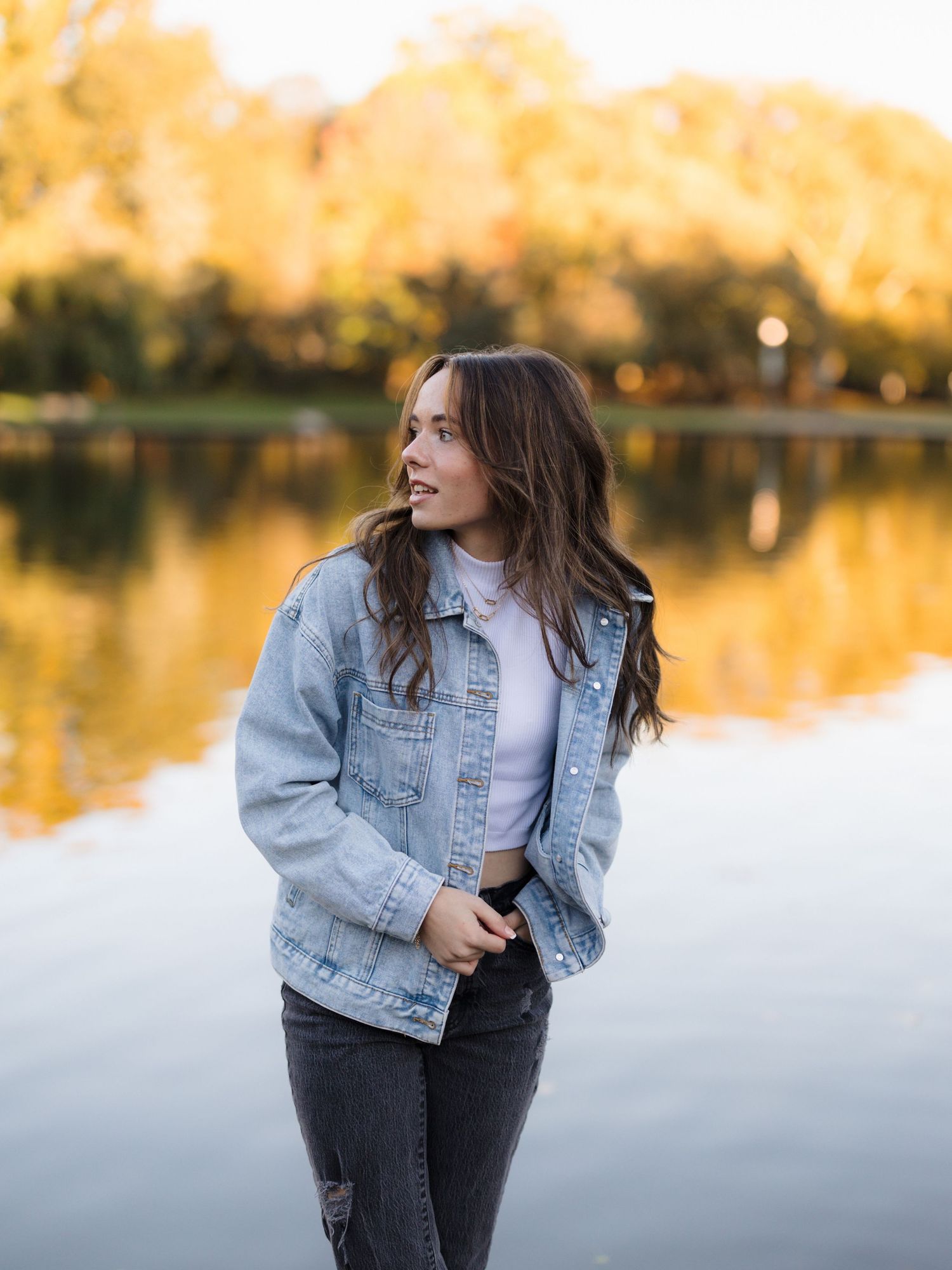 Young woman with long brown hair in a denim jacket and gray jeans stands by a calm lake with golden autumn trees reflected in the water.
