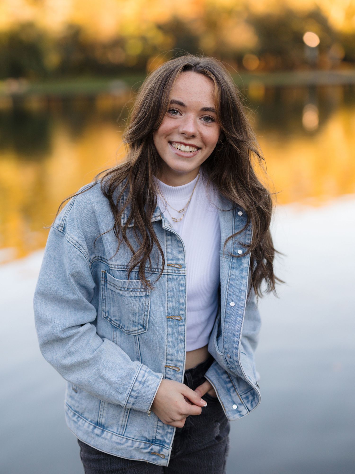 Smiling young woman with long brown hair wearing a denim jacket and white top stands by a calm lake at golden hour.