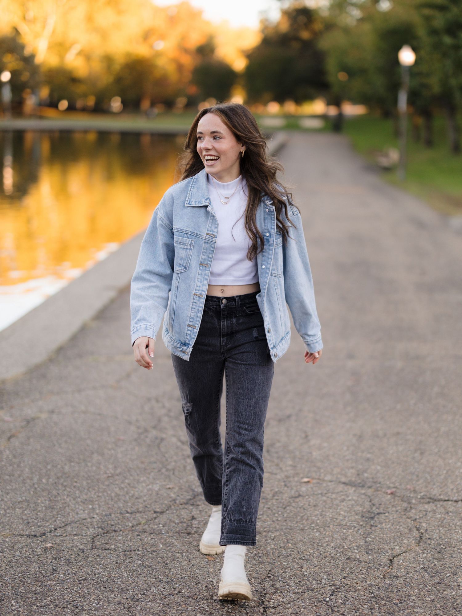 Smiling woman in a light denim jacket and black jeans walks along a paved path beside a calm lake at golden hour.