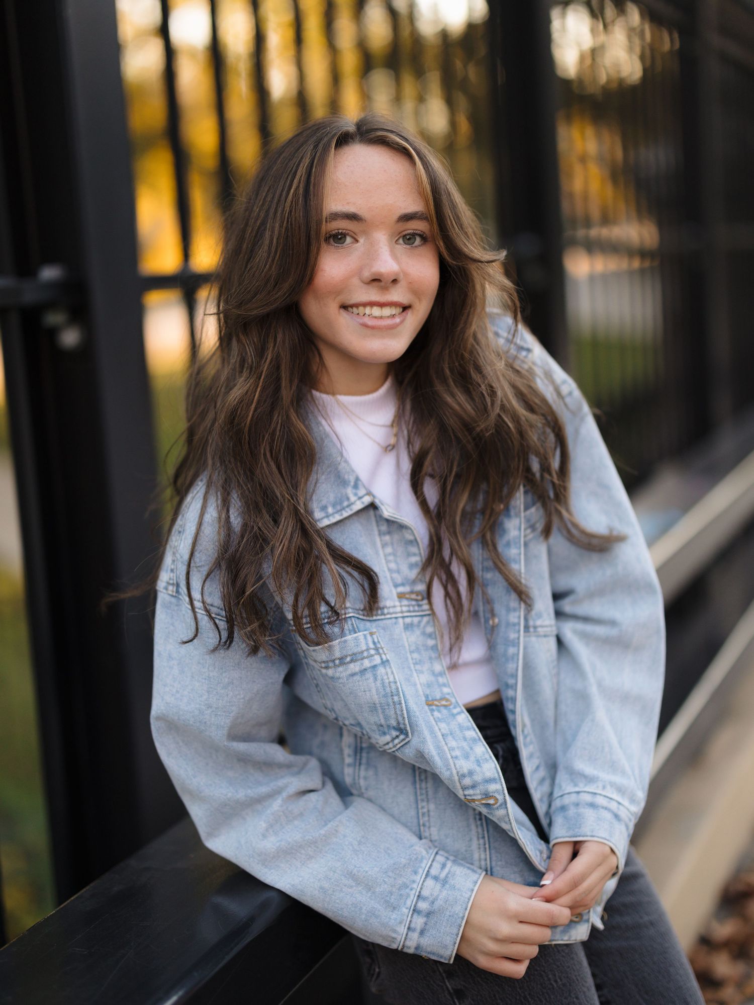 Smiling young woman with long wavy brown hair wearing a light denim jacket over a white top, sitting by a black metal fence outdoors.