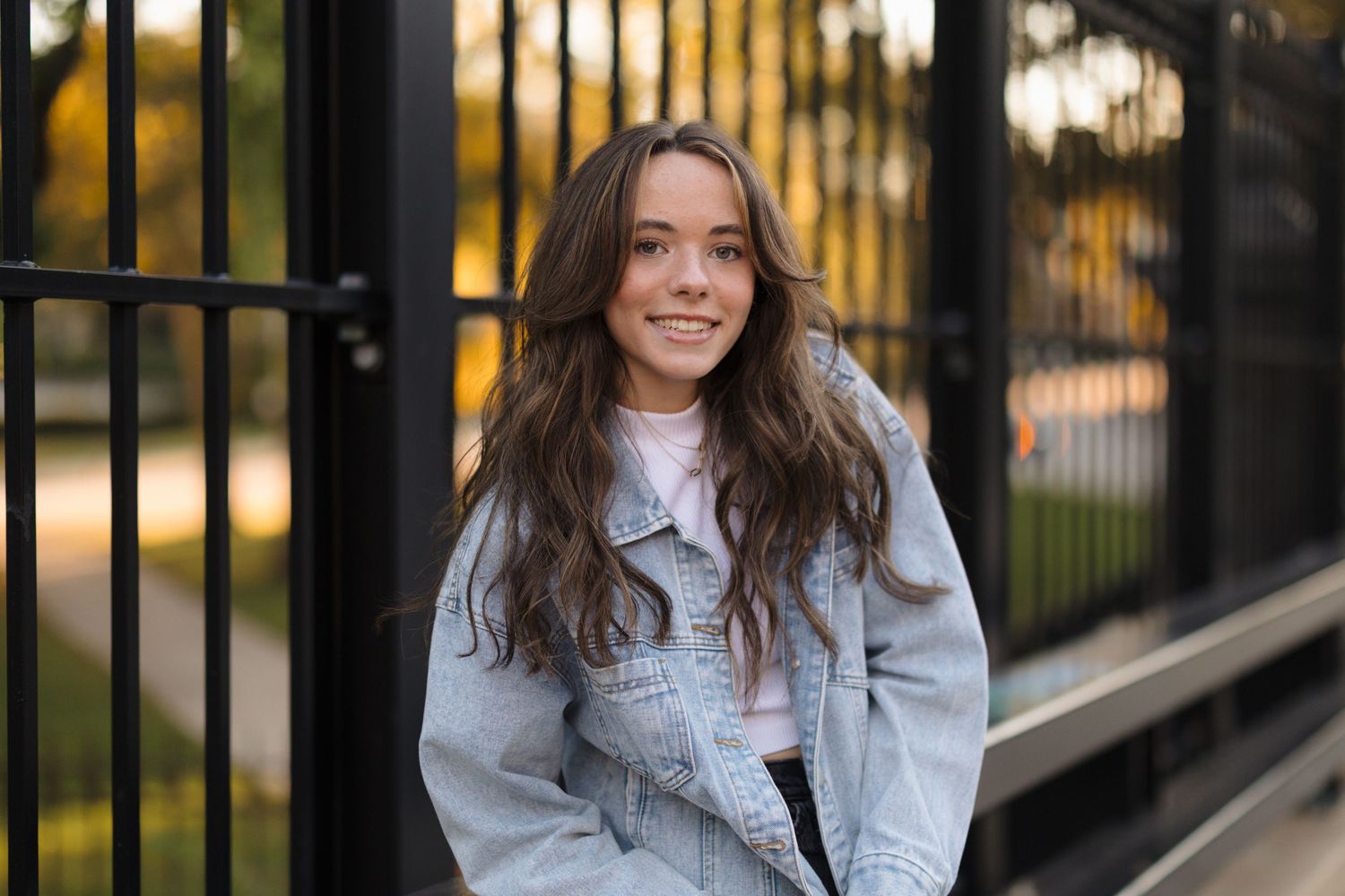 Smiling young woman with long wavy brown hair wearing a light denim jacket stands beside a black metal fence in warm golden-hour light.
