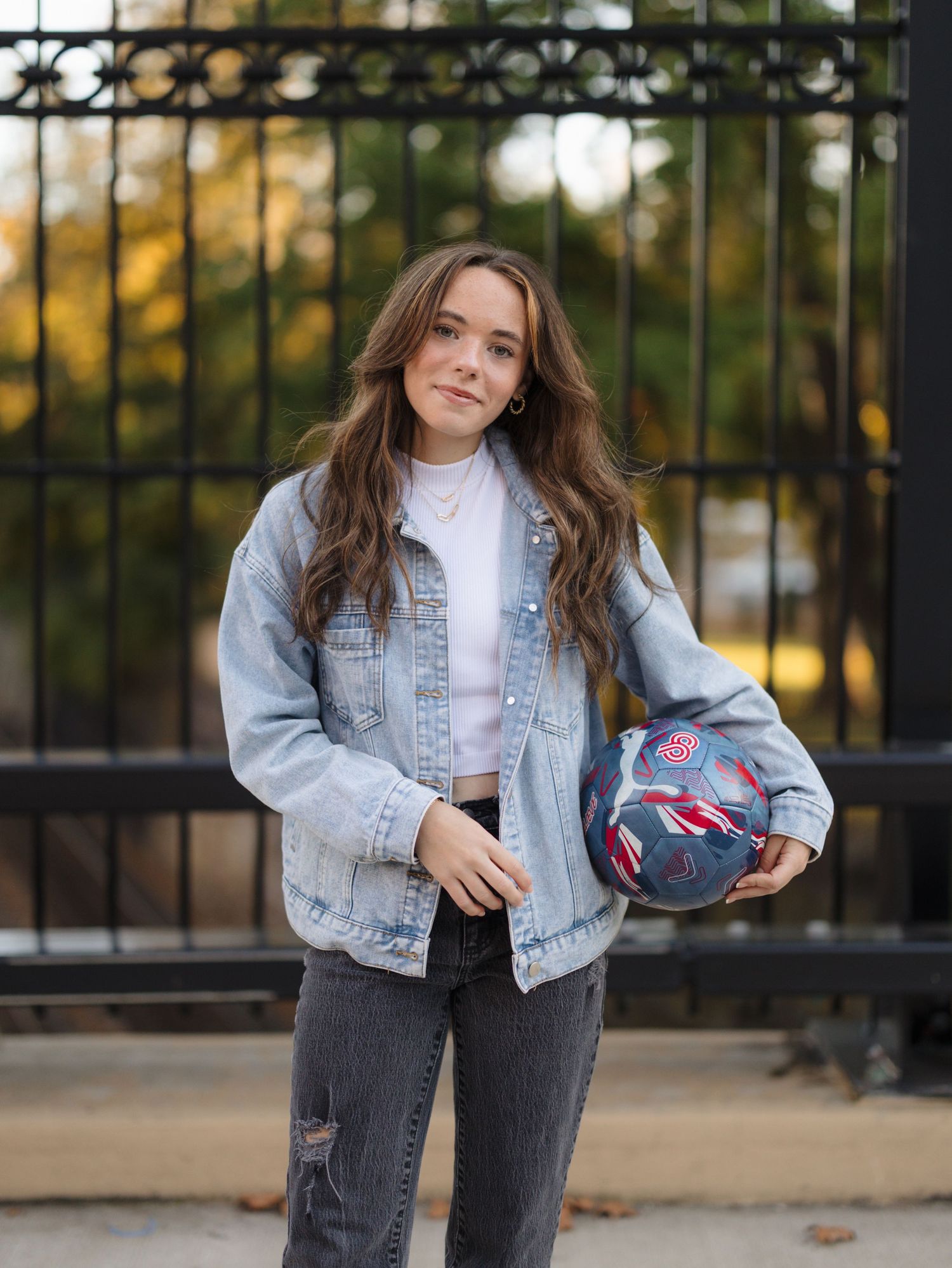 Smiling young woman with wavy hair wears a light denim jacket and black jeans, holding a patterned ball in front of a black iron fence.