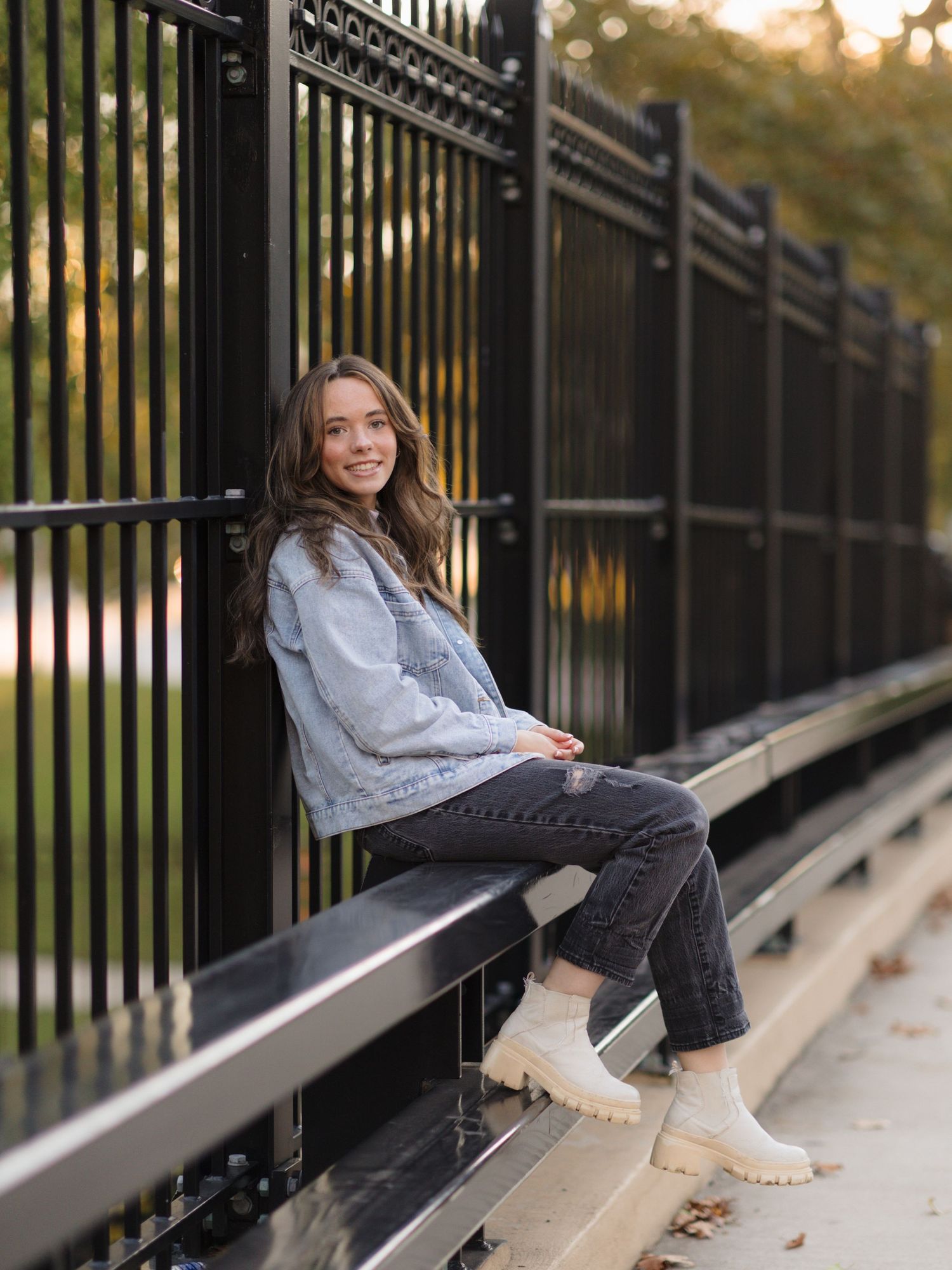 Smiling young woman with long brown hair in a denim jacket and gray jeans sits on a ledge beside a black iron fence outdoors.
