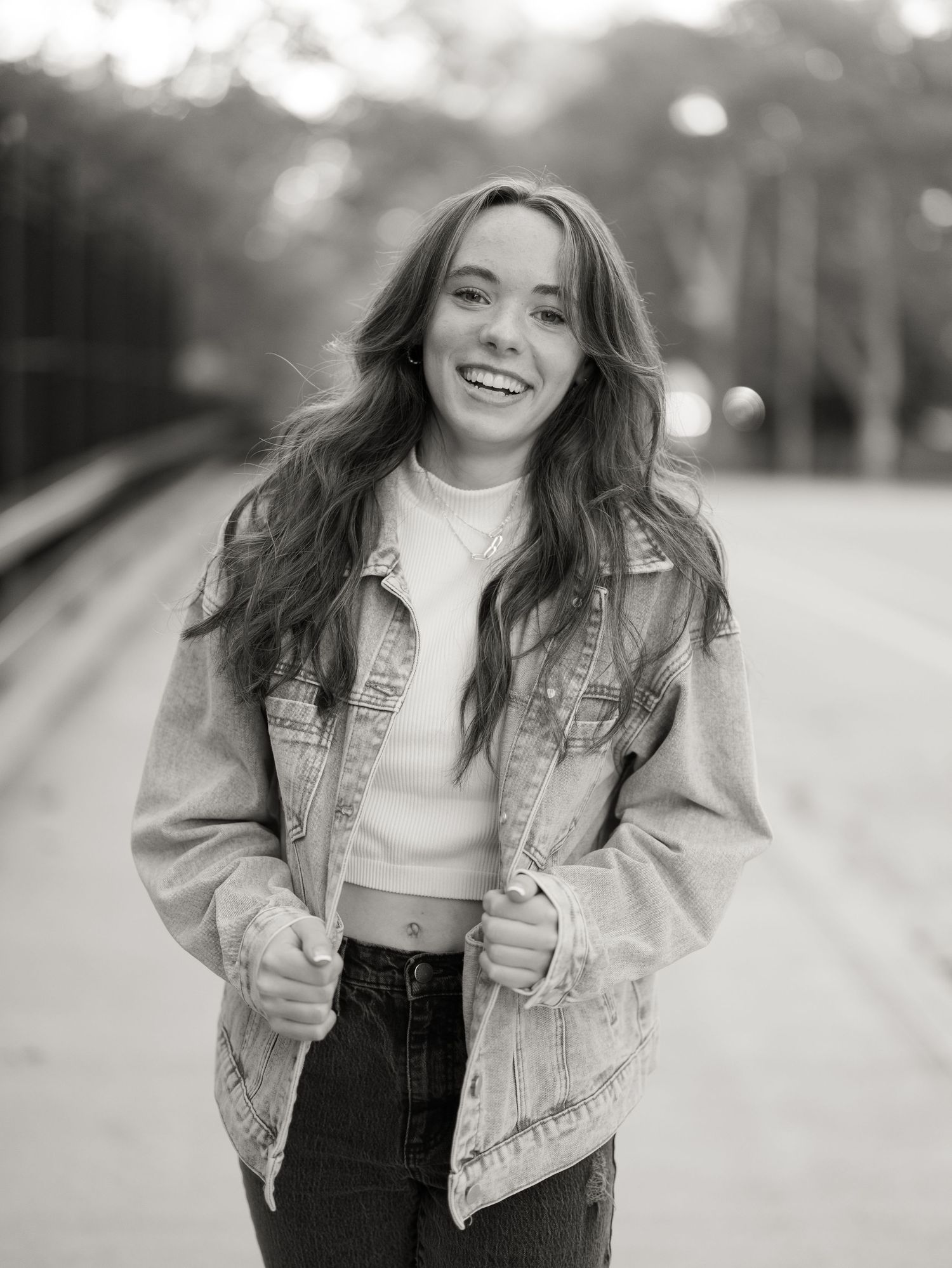 Smiling young woman with long wavy hair wearing a denim jacket over a cropped top, posing outdoors in a black and white portrait.