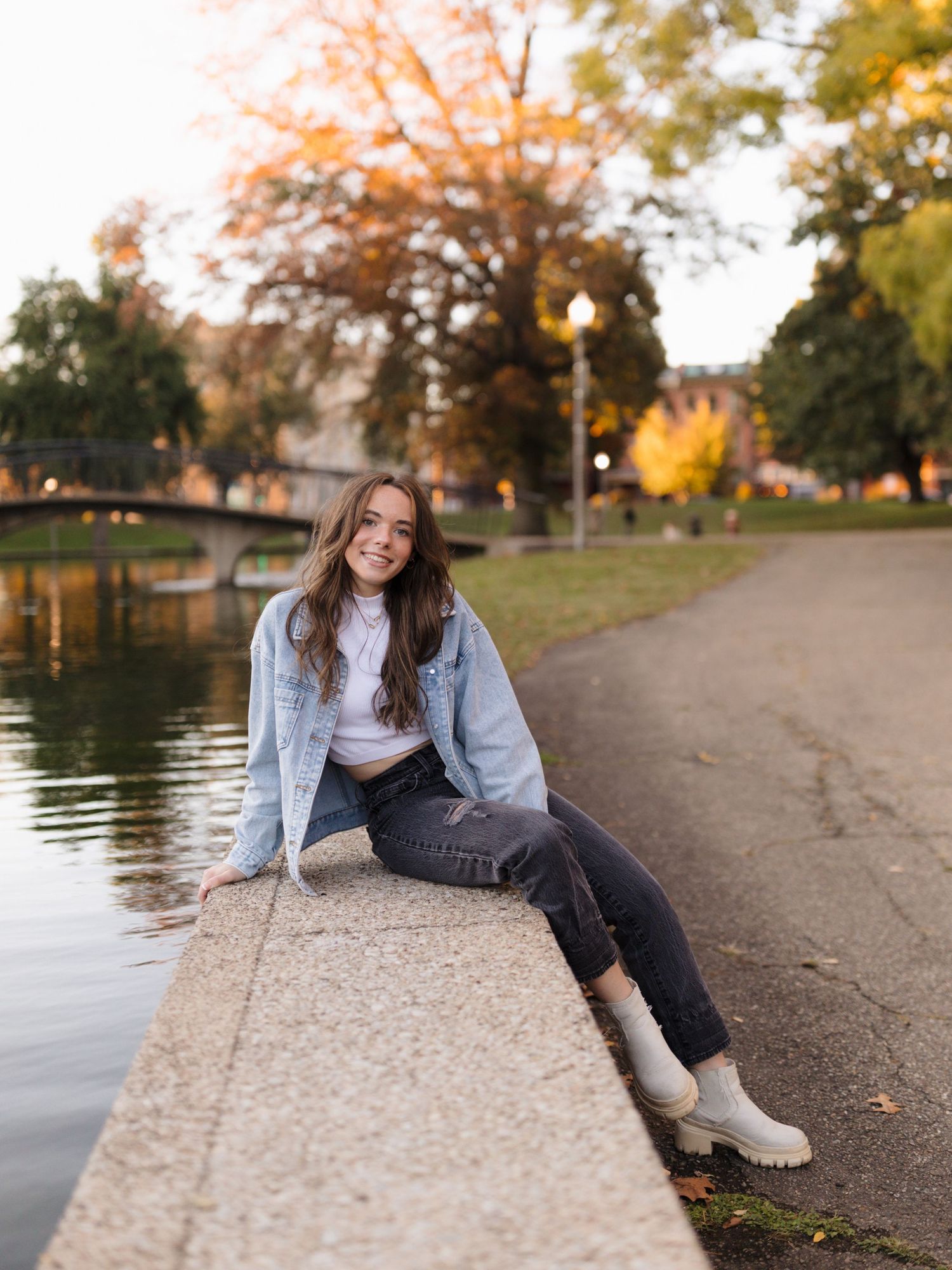 Smiling young woman in a denim jacket and white boots sits on a stone ledge beside a calm pond with colorful autumn trees nearby.