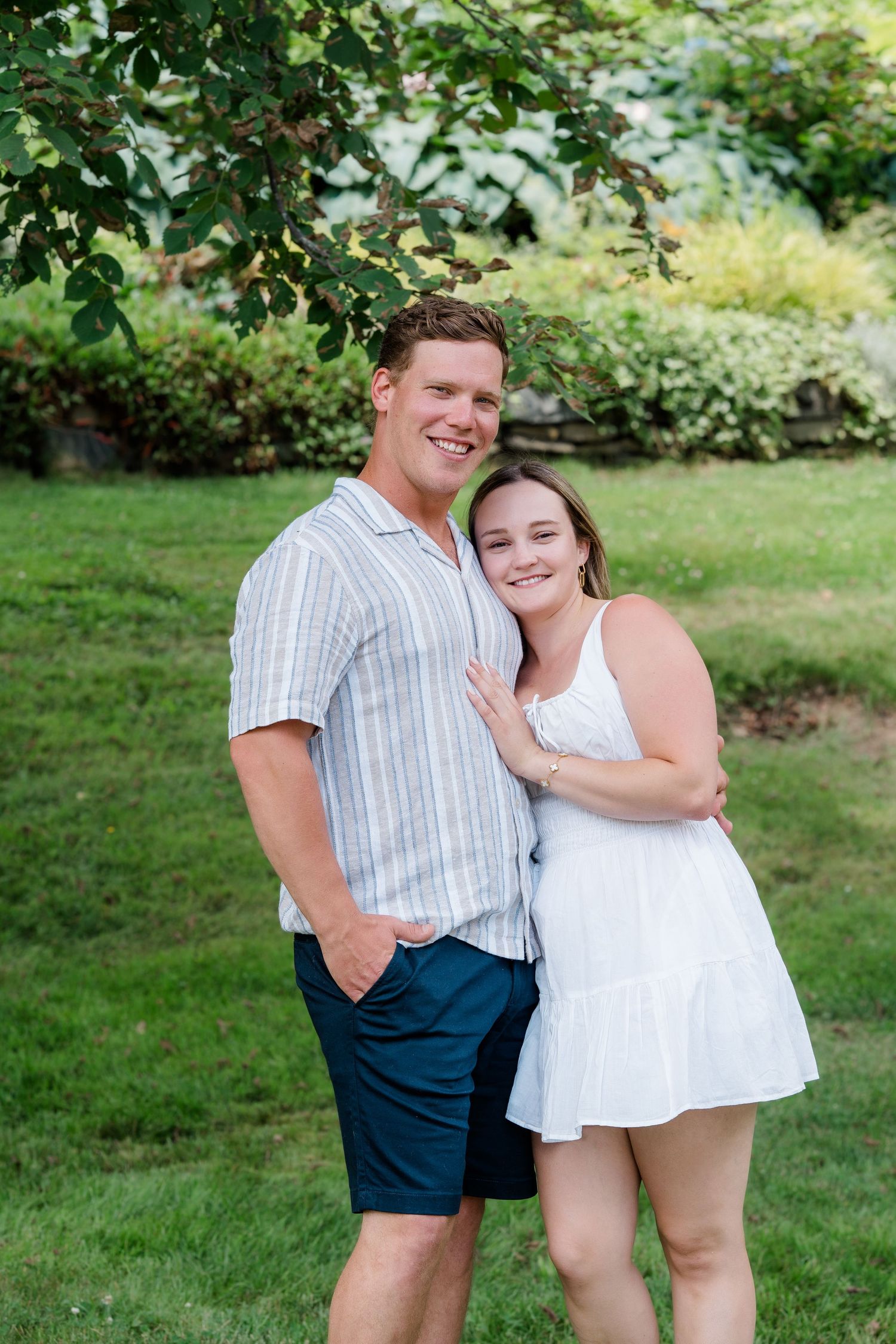 Smiling couple embraces outdoors in a green garden, man in striped shirt and woman in white dress at Dartmouth Commons.