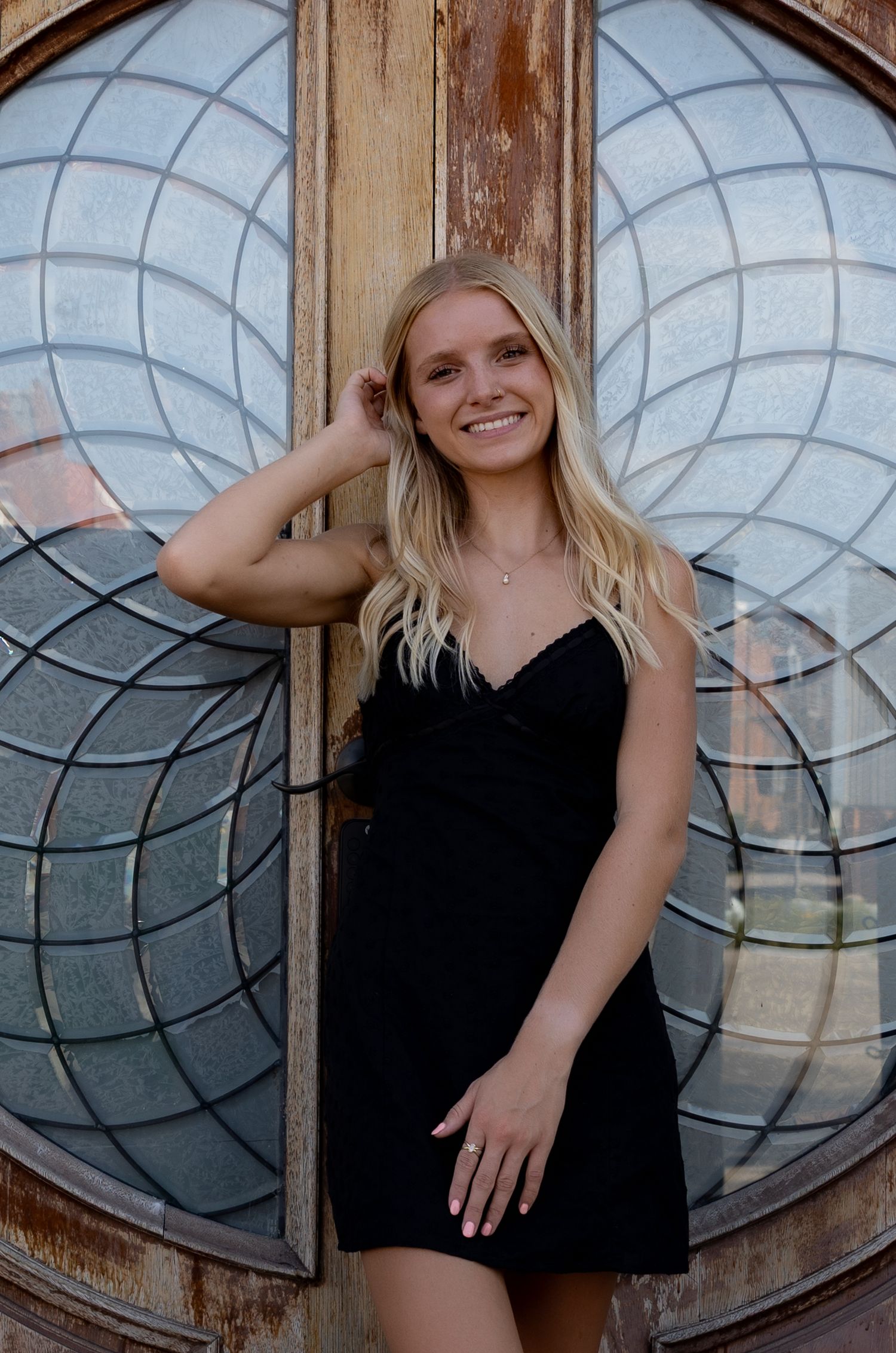 Smiling blonde woman in black mini dress posing in front of ornate stained glass door.