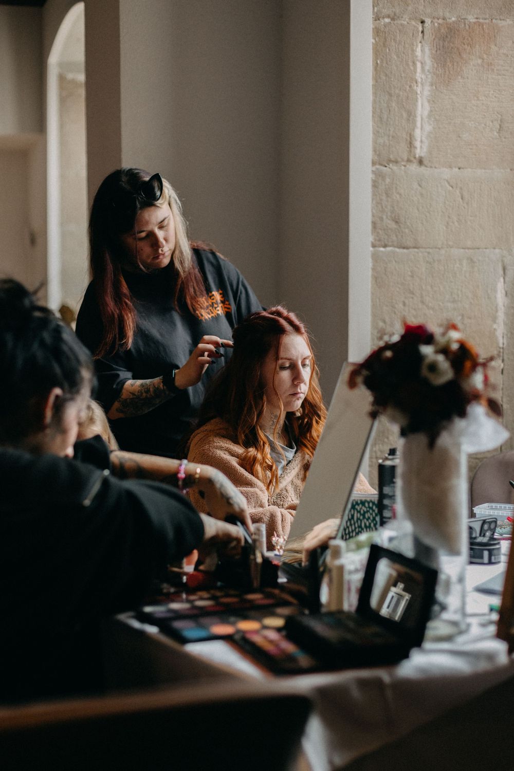 Makeup artist applies cosmetics to a woman with auburn hair at a table filled with eyeshadow palettes and a floral arrangement.