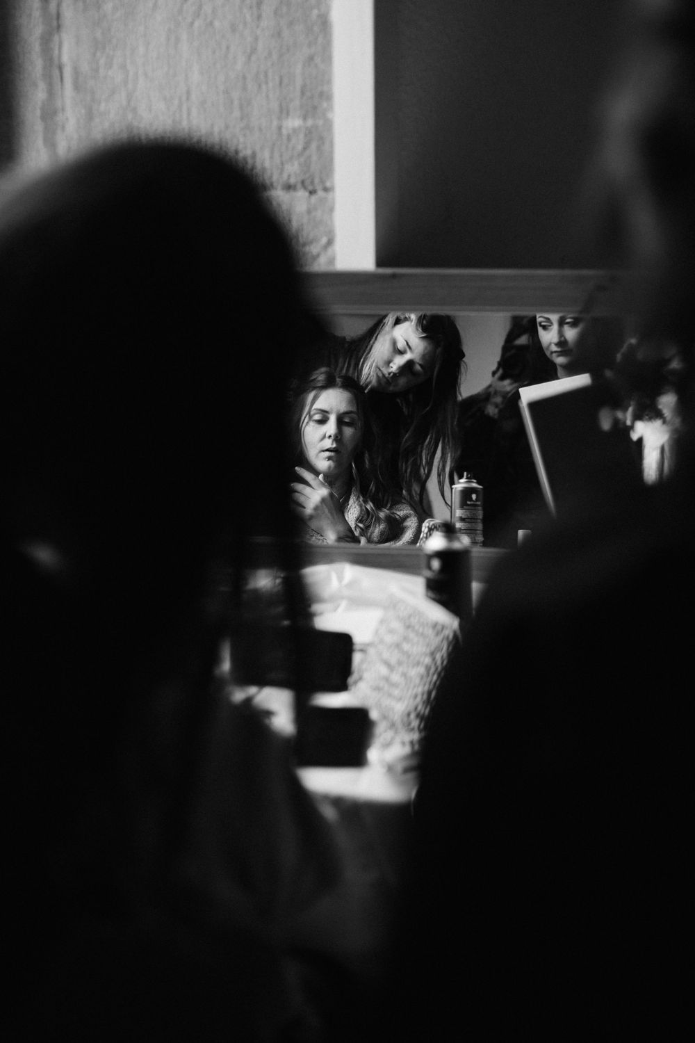 Black and white mirror reflection of a bride getting ready, with bridesmaids gathered close behind her as she prepares.