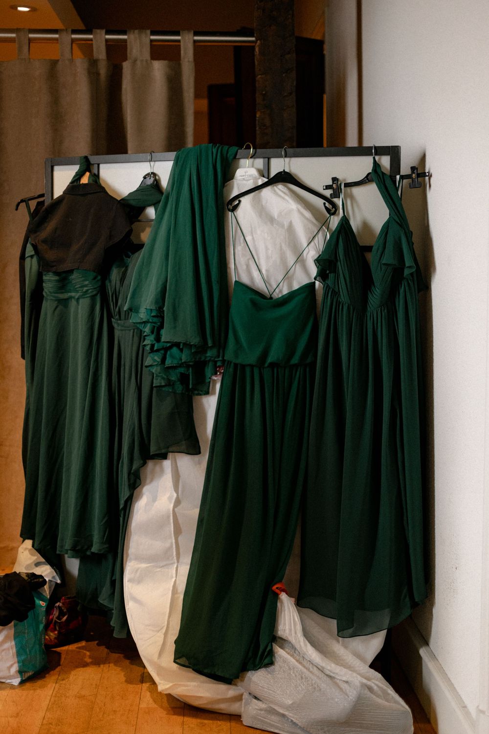 Emerald green bridesmaid dresses and a white wedding gown hanging on a rack in a wedding preparation room.