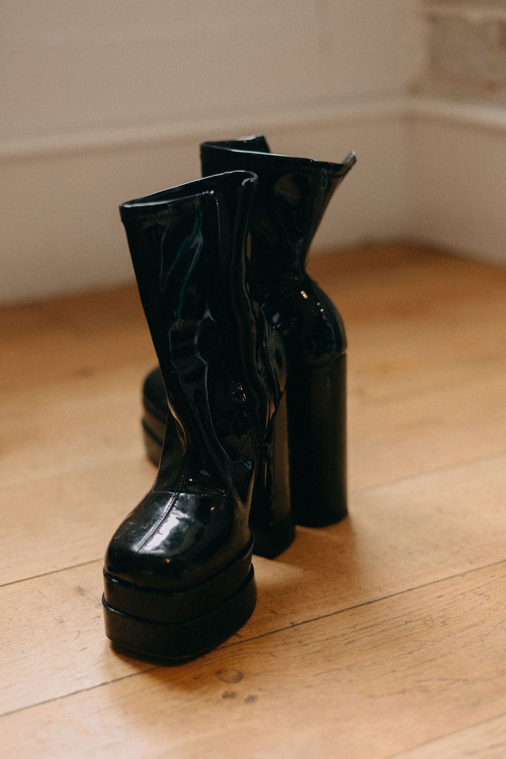 A pair of glossy black platform ankle boots with chunky heels stand on a light wooden floor.