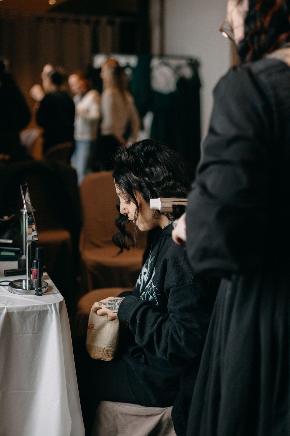 A woman in a black sweatshirt sits at a table while a stylist works on her hair during wedding preparations.