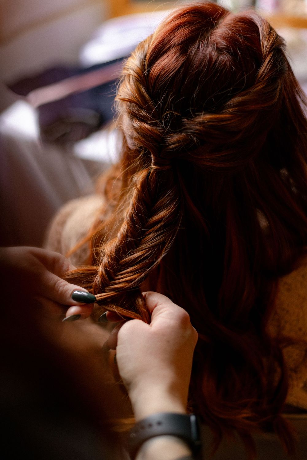 Close-up of a hairstylist weaving a loose fishtail braid into long wavy auburn hair for a wedding hairstyle.