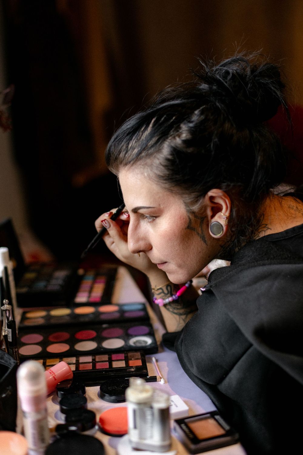 Woman with dark hair in a bun applies eyeshadow in front of a mirror beside colorful makeup palettes and products.