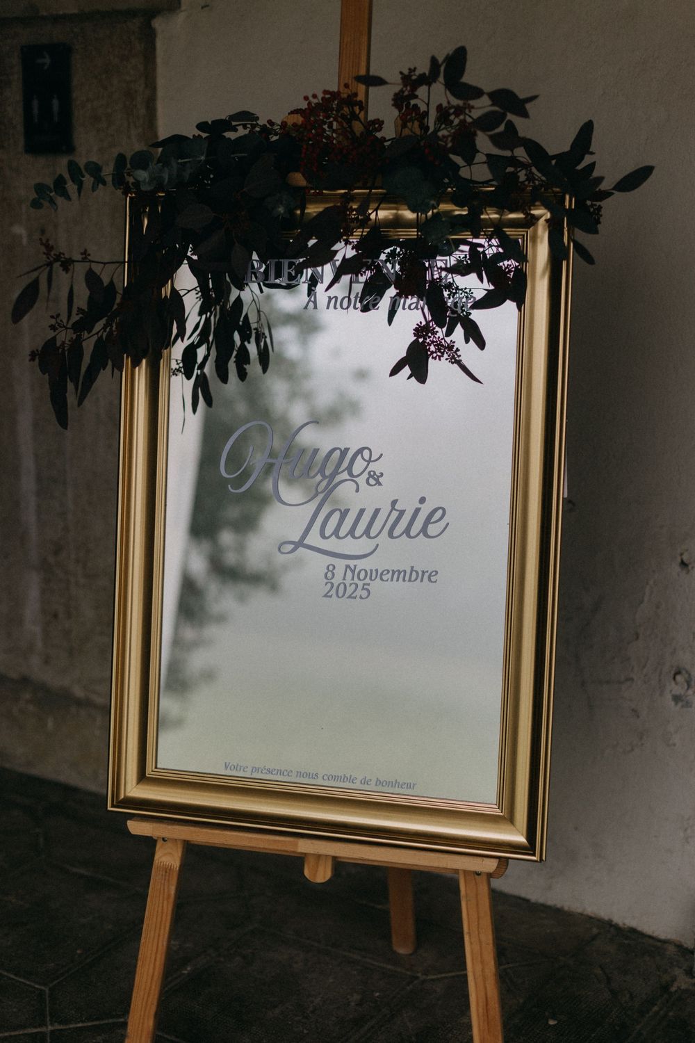 Gold-framed wedding welcome mirror sign for Hugo and Laurie, dated 8 November 2025, topped with dark eucalyptus foliage on an easel.